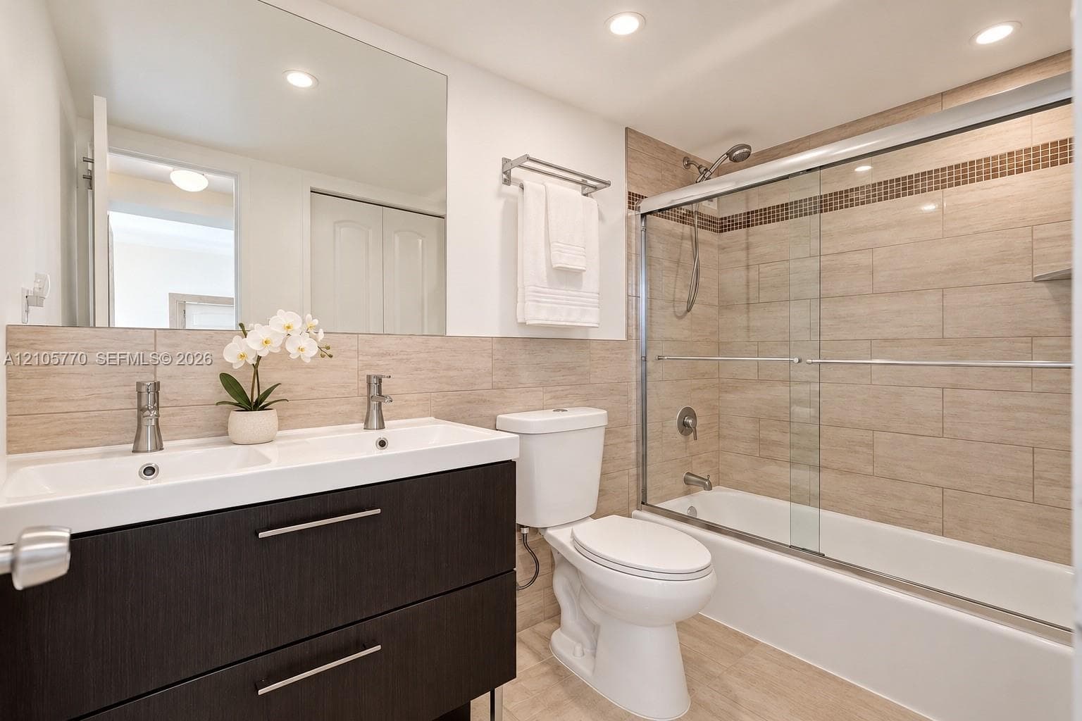 Dark wood-finish double vanity with dual faucets and integrated sink, complemented by a large wall-mounted mirror, recessed lighting, and a bathtub with glass sliding doors and full-height tile surround