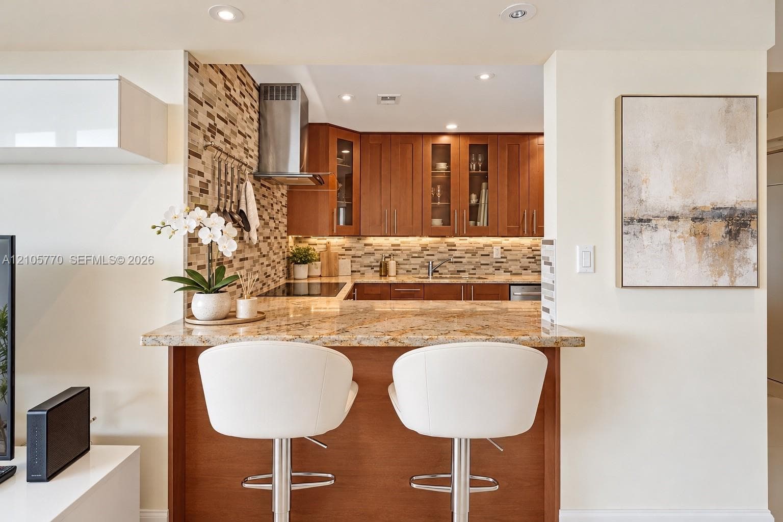 Kitchen featuring a stone-surface breakfast bar, wood cabinetry with glass-front uppers, mosaic tile backsplash, and stainless steel range hood