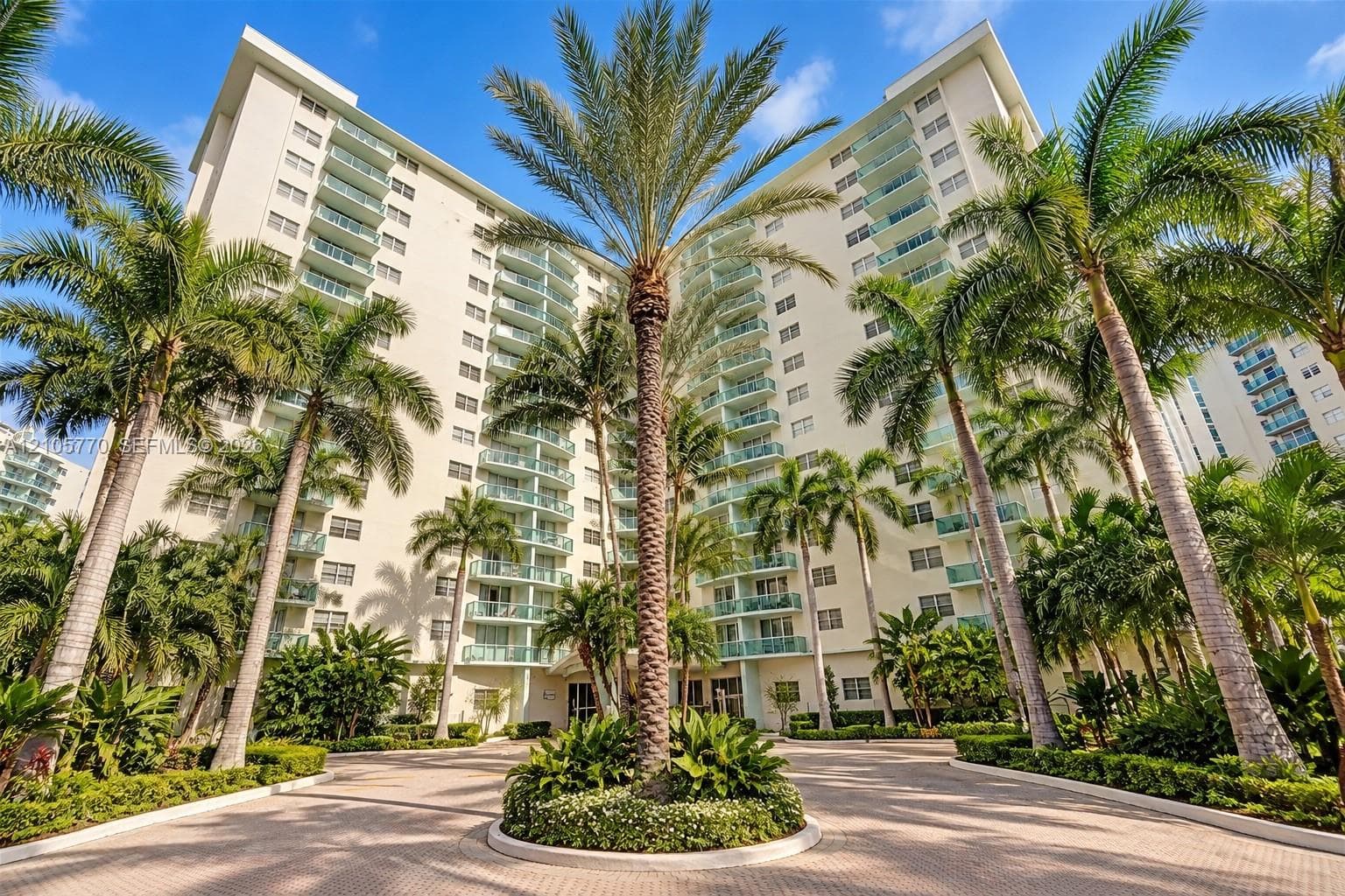 High-rise residential exterior featuring a paved circular driveway, mature palm trees, multi-level private balconies with glass railings, and expansive stucco facade