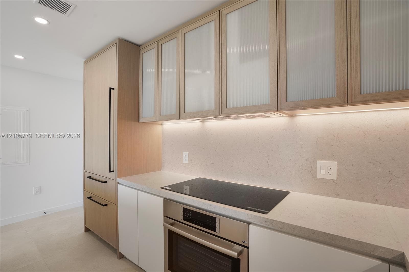 Custom cabinetry featuring light wood finish and reeded glass upper cabinet doors, integrated refrigeration, stone-finish countertops, and an induction cooktop with stainless steel wall oven