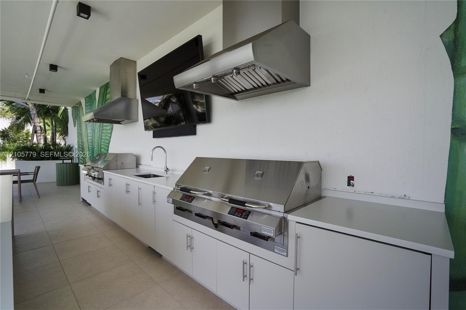 Outdoor kitchen featuring dual stainless steel ventilation hoods, built-in stainless steel grill, integrated sink with gooseneck faucet, and white cabinetry with solid-surface countertops