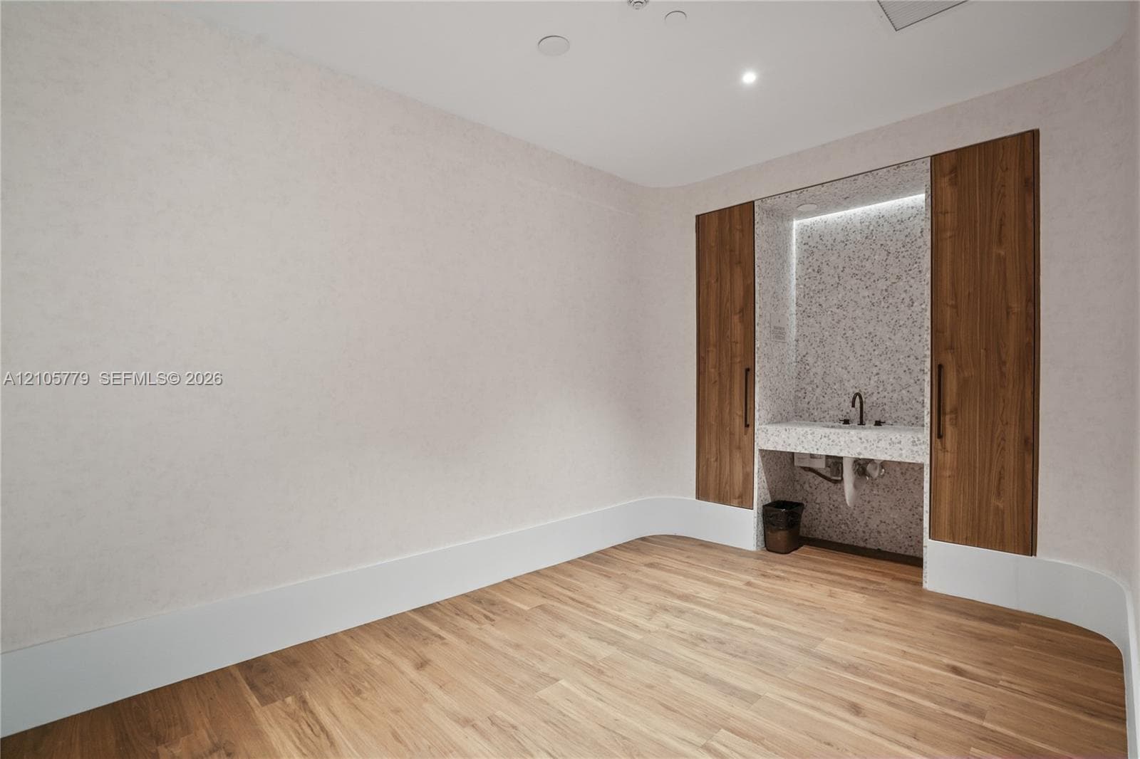 Room featuring wood-finish flooring, curved wall architecture, built-in floor-to-ceiling wood cabinetry, and a stone-surround recessed vanity with integrated LED lighting and a matte black faucet. Private massage room.