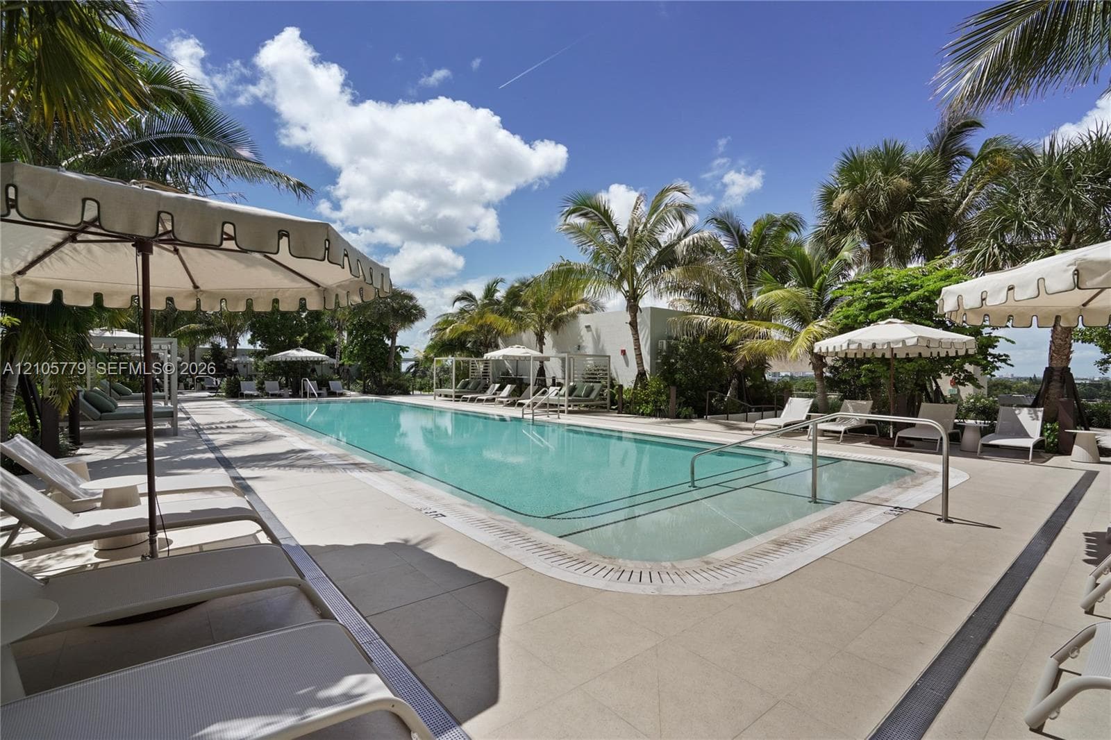 Rectangular swimming pool featuring stone-tile decking, metal handrails, integrated pool steps, and perimeter landscaping with mature palms