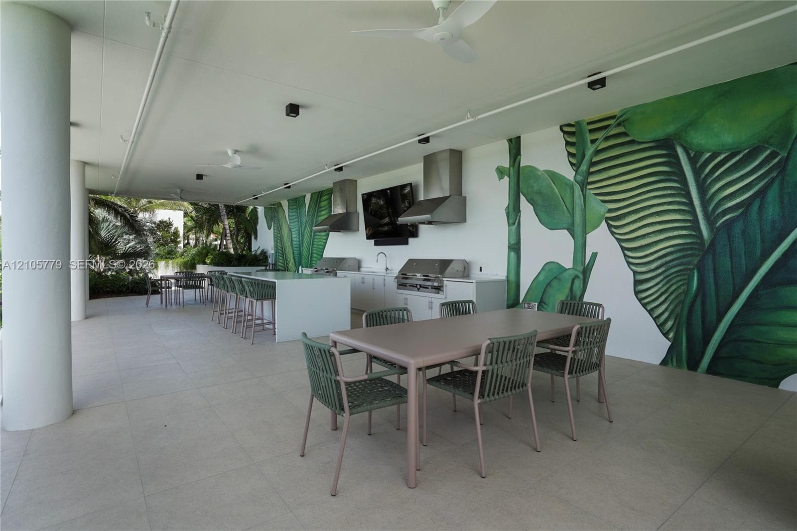 Covered outdoor kitchen featuring a large stone-finish island with integrated seating, built-in stainless steel appliances, dual vent hoods, and oversized wall mural detailing