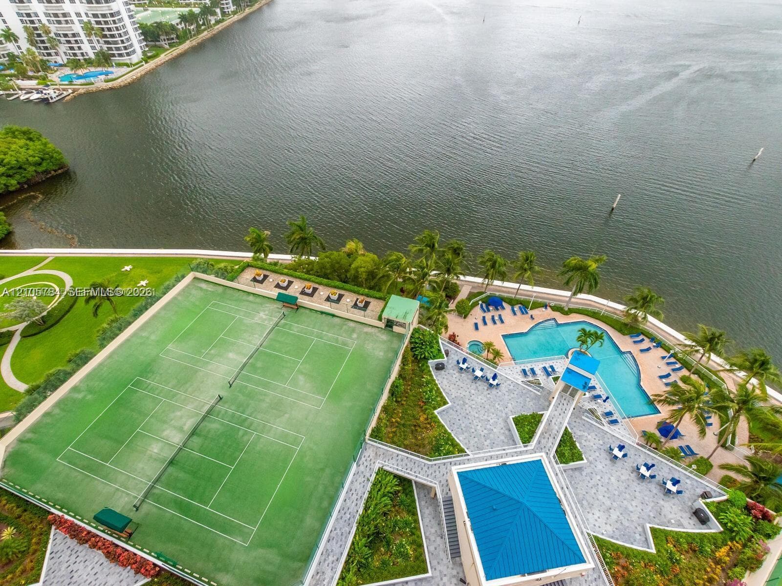 Aerial perspective of property grounds featuring a tennis court, swimming pool with spa, paved patio area, and mature palm trees