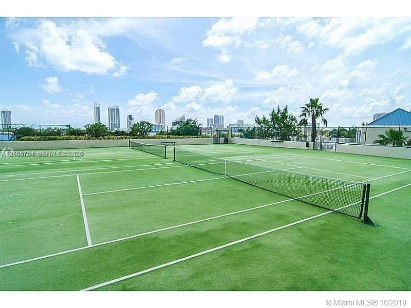 Tennis court featuring a specialized synthetic surface, full-length perimeter fencing, a central net system, and urban skyline backdrop