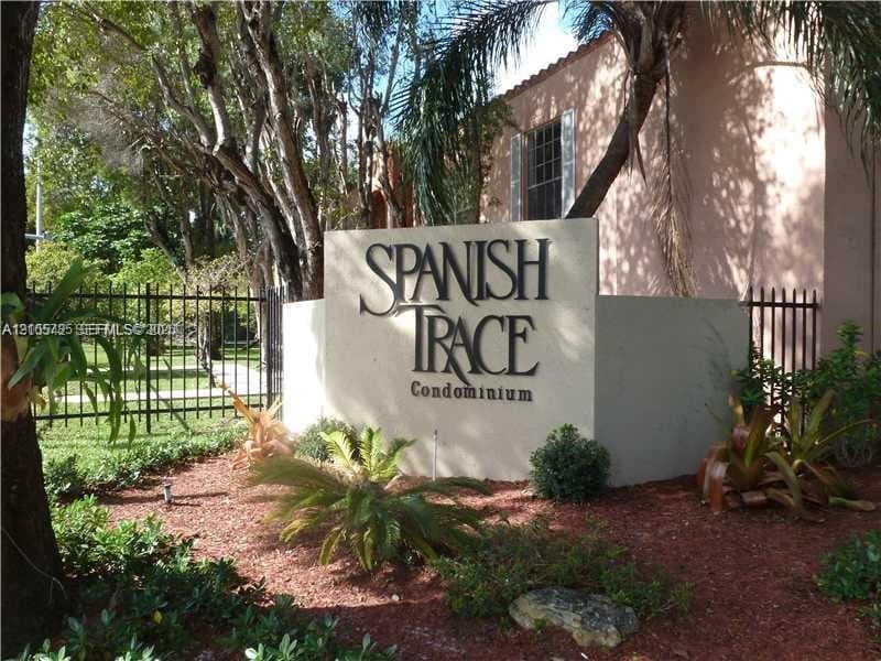 Stucco monument sign featuring dimensional lettering, surrounded by mature trees, ornamental shrubs, mulch landscaping, and black metal perimeter fencing