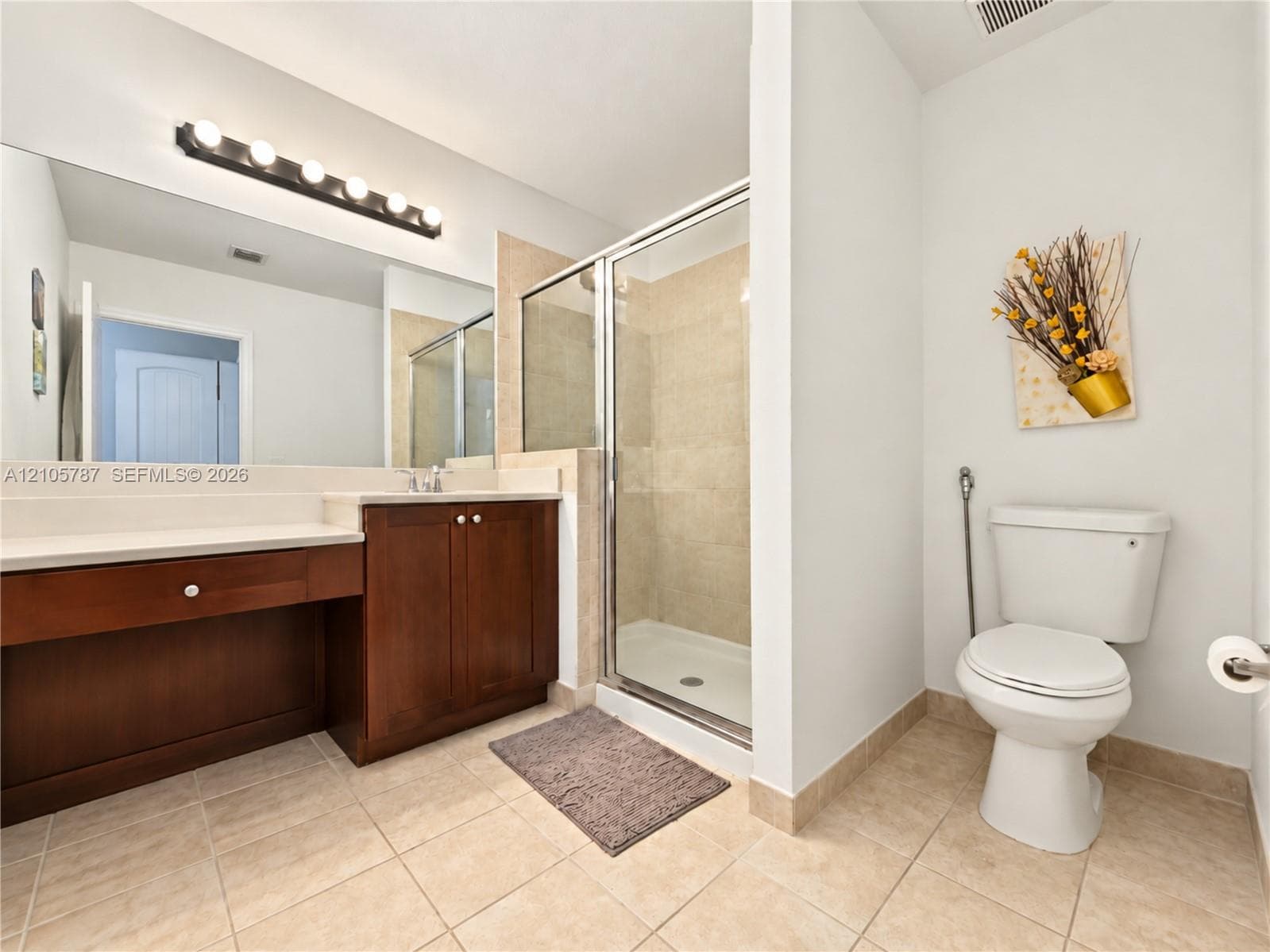 Bathroom featuring a wood-finish vanity with a light-toned countertop, a large wall-mounted mirror with multi-bulb light fixture, a glass-enclosed shower with tile surround, and tile flooring