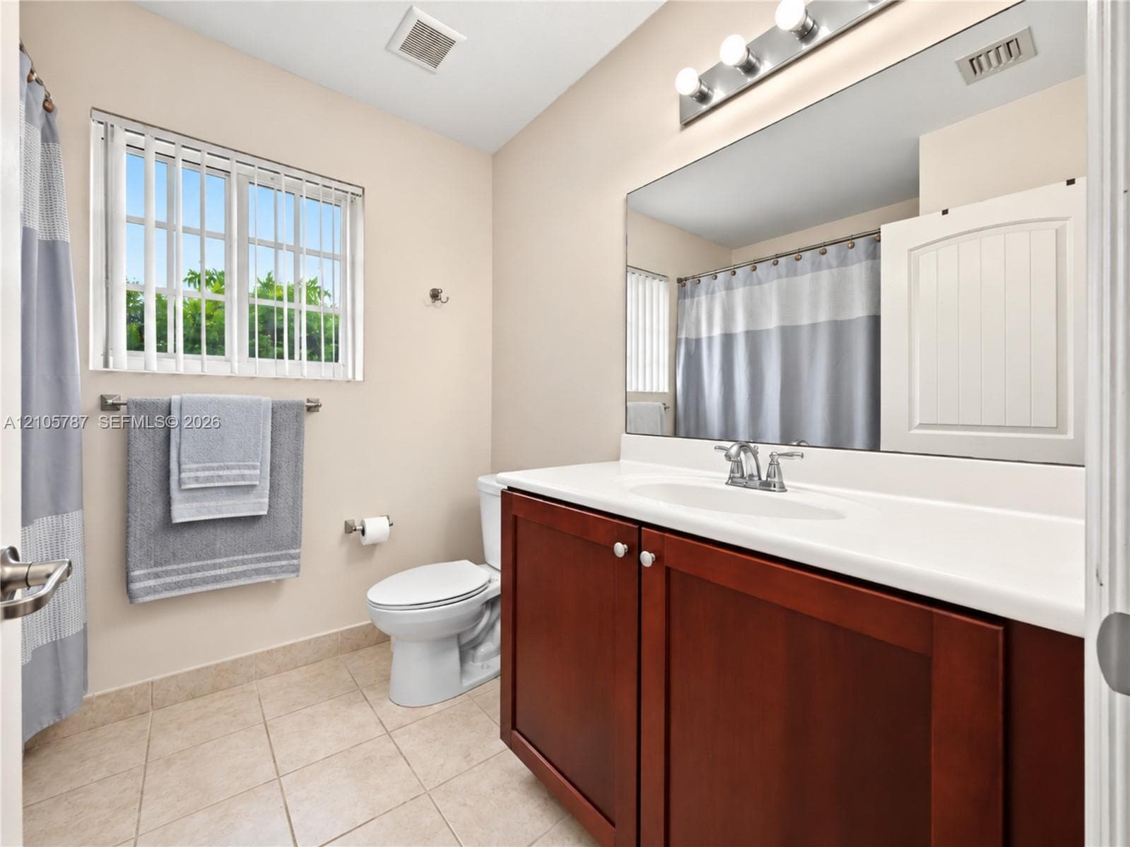 Bathroom featuring wood-finish vanity cabinetry, solid-surface countertop with an integrated sink, full-width vanity mirror, four-bulb fixture, and tile flooring