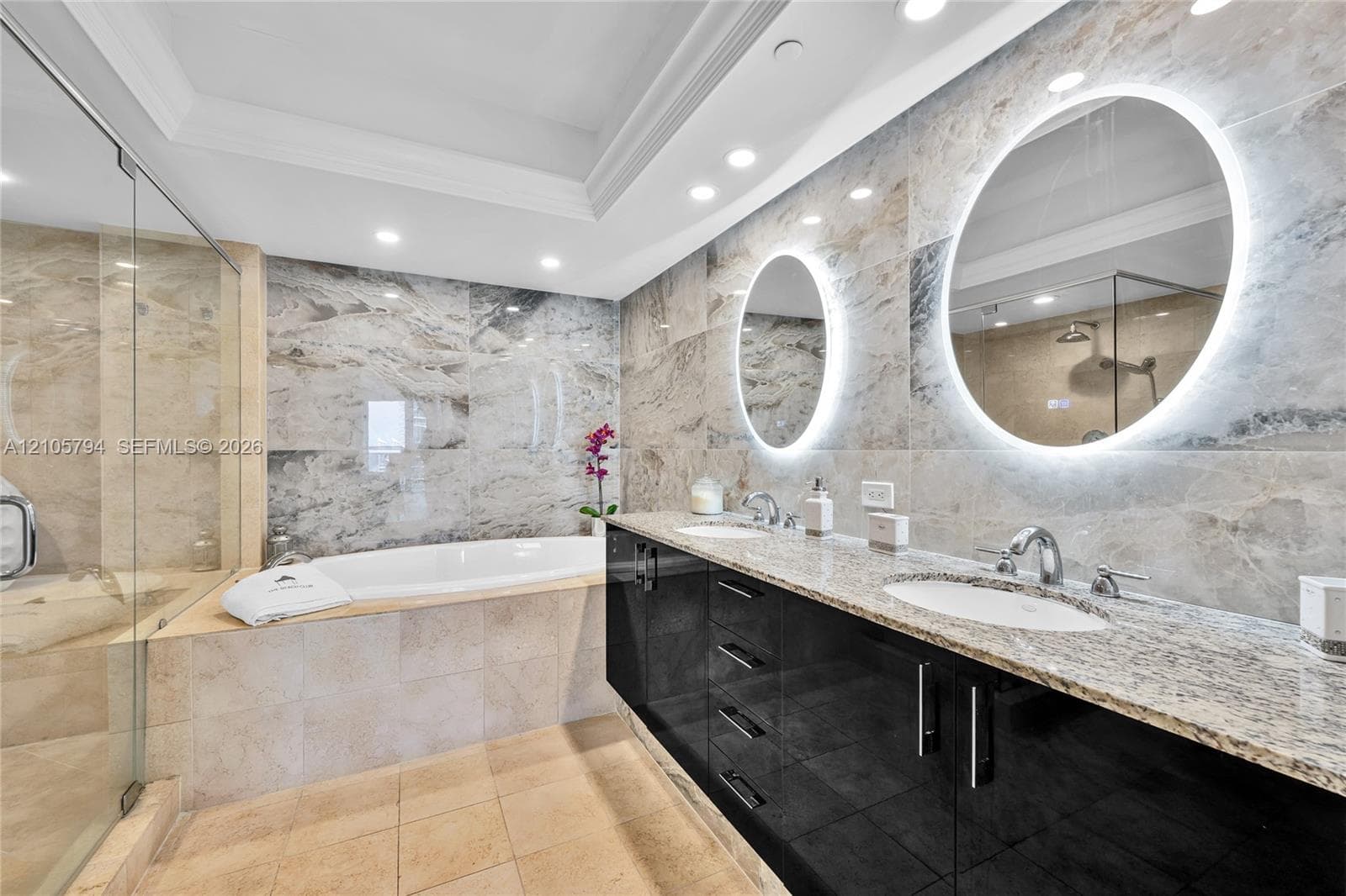 Double vanity with high-gloss cabinetry and stone countertops, oval LED-backlit mirrors, soaking tub with tile surround, and walk-in glass-enclosed shower