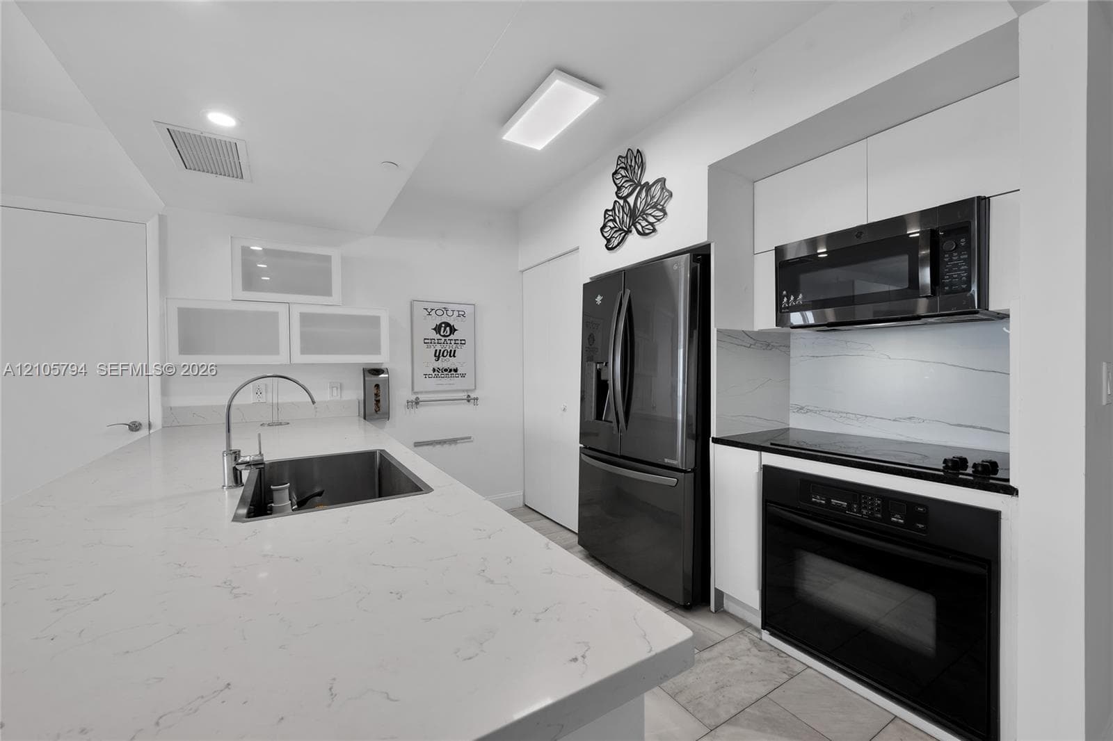 Kitchen featuring a central island with a stone-finish countertop and undermount sink, white cabinetry with frosted glass accents, stone-finish backsplash, built-in stainless steel microwave and oven, and dark-toned full-size refrigerator