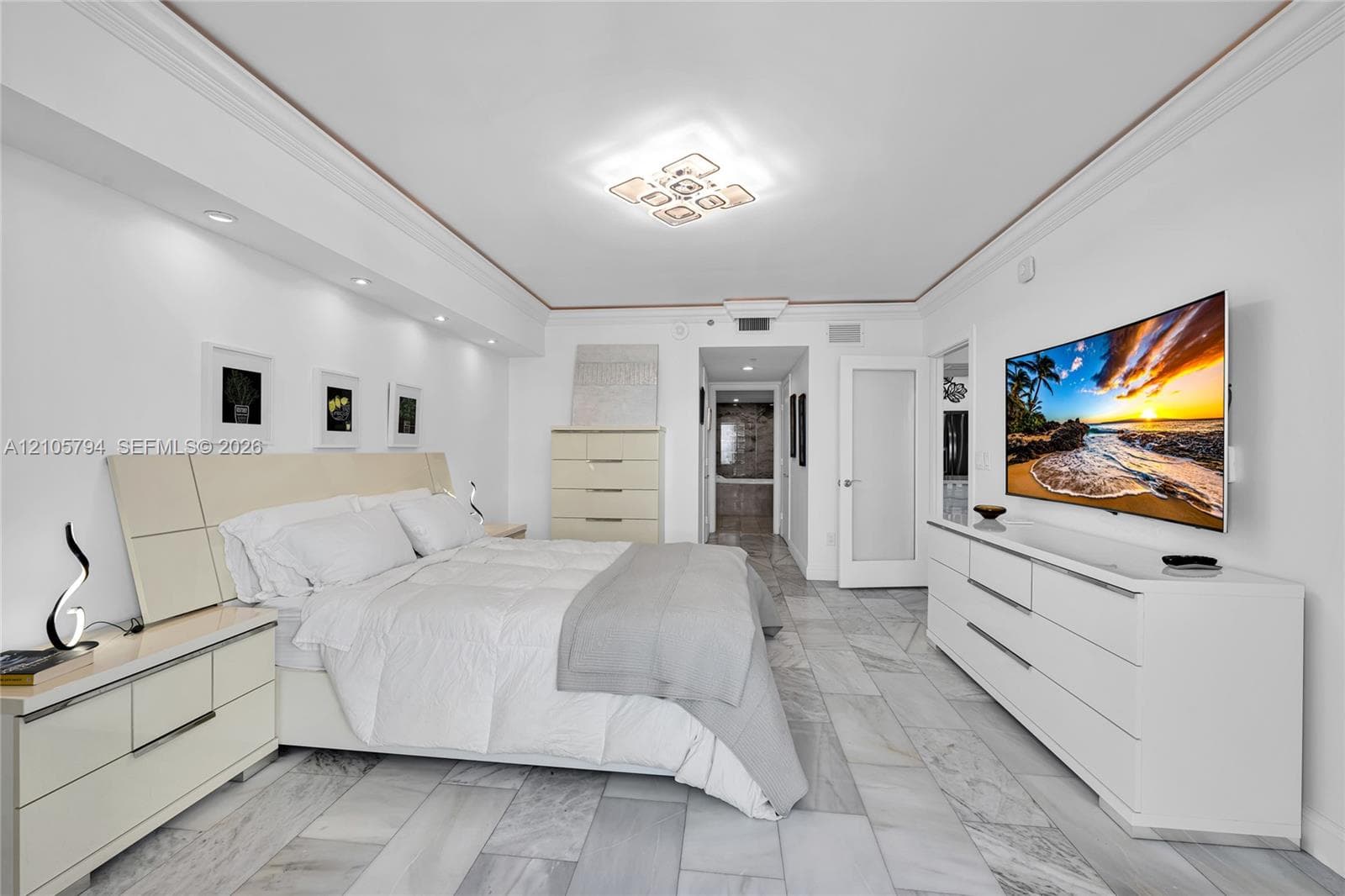 Neutral-toned interior featuring large-format stone-finish floor tile, recessed ceiling lighting, crown molding, and a dedicated wall-mounted lighting feature above the headboard area