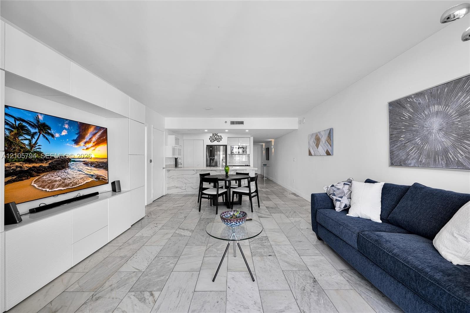 Open-concept living space featuring stone-style tile flooring, floor-to-ceiling built-in white cabinetry, recessed ceiling lighting, and a breakfast bar with stone-style countertops