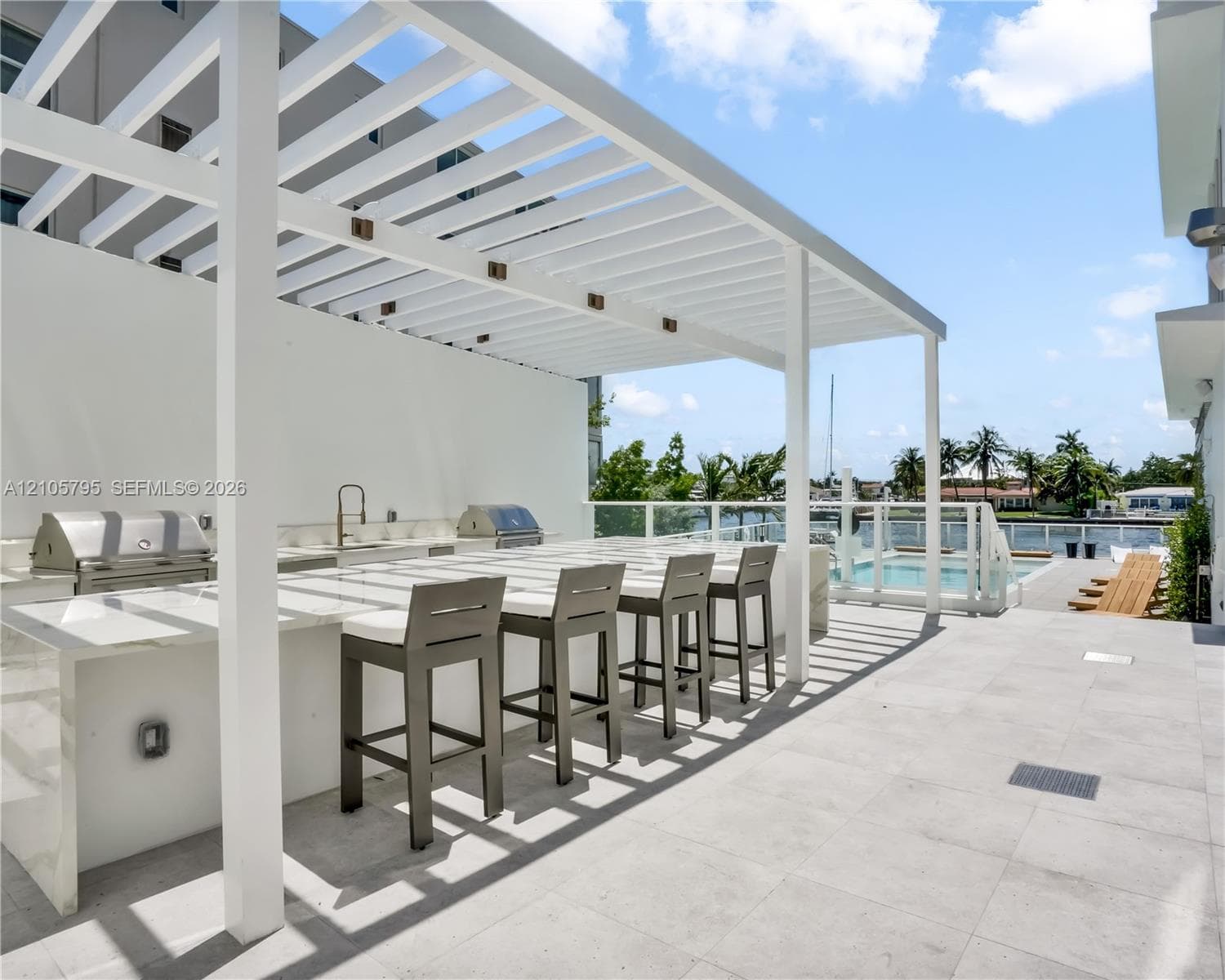 Outdoor kitchen featuring a stone-topped island with bar seating, integrated stainless steel grill, pergola covering, and concrete tile flooring