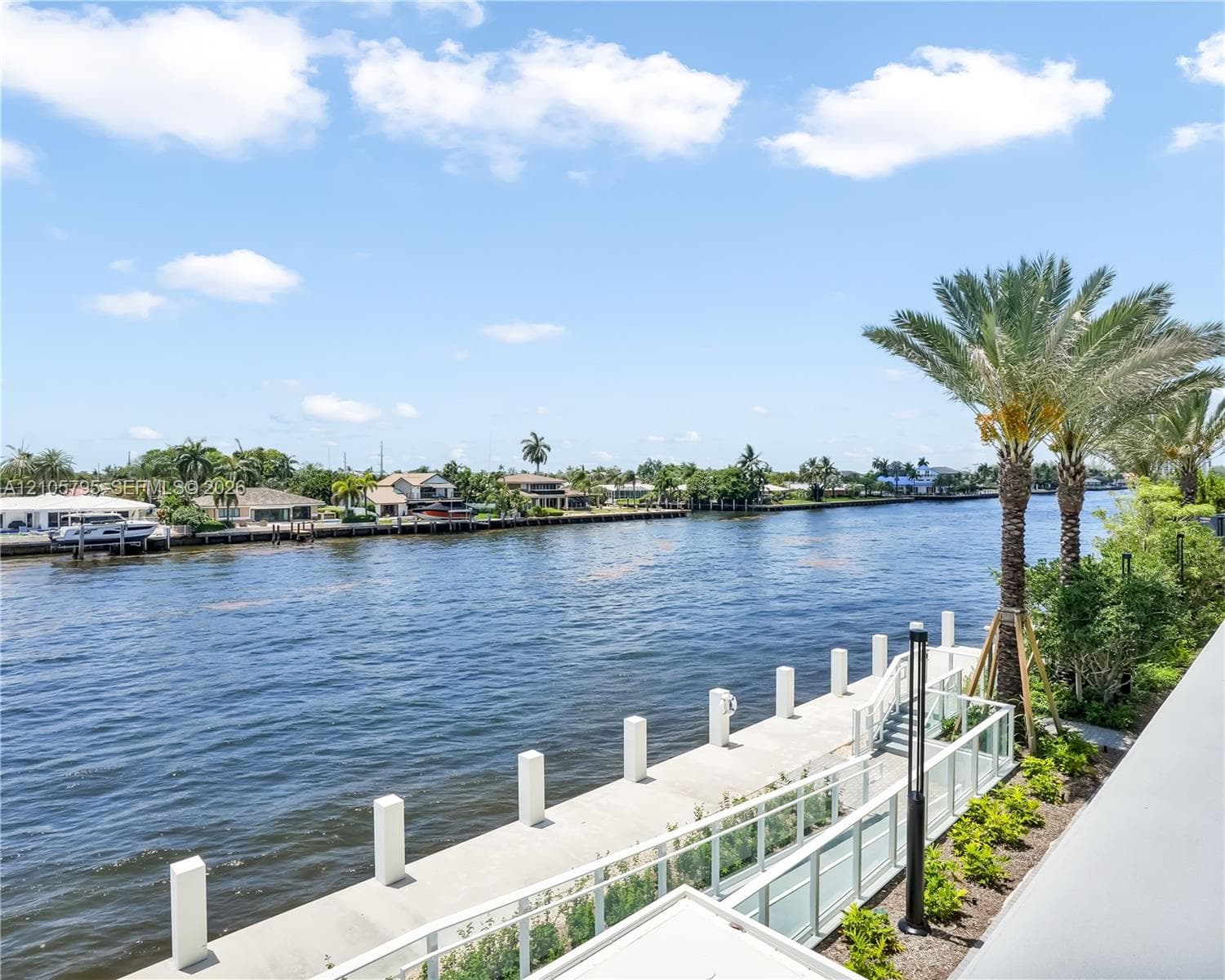 Waterfront property featuring a concrete seawall, glass panel railing, paved walkway, and mature palm trees