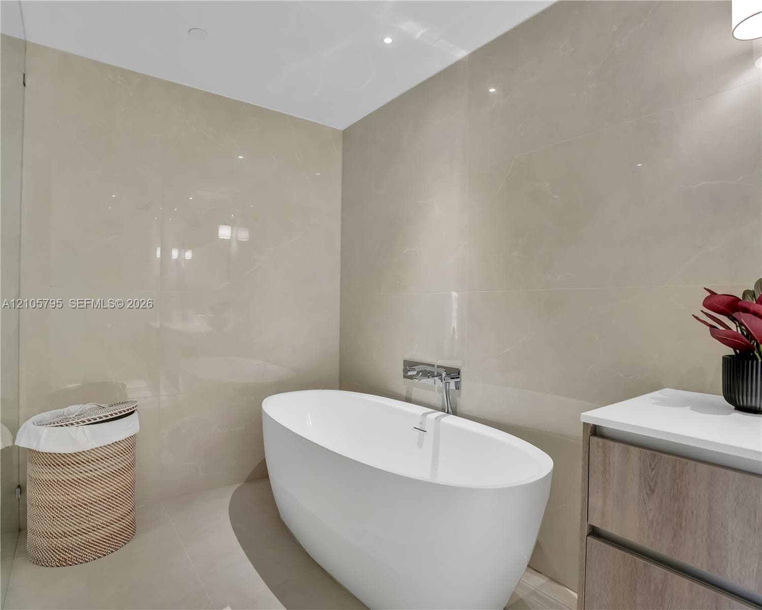 Freestanding white soaking tub with wall-mounted polished chrome faucet, floor-to-ceiling large-format tile wall cladding, and wood-finish vanity cabinet with a white countertop