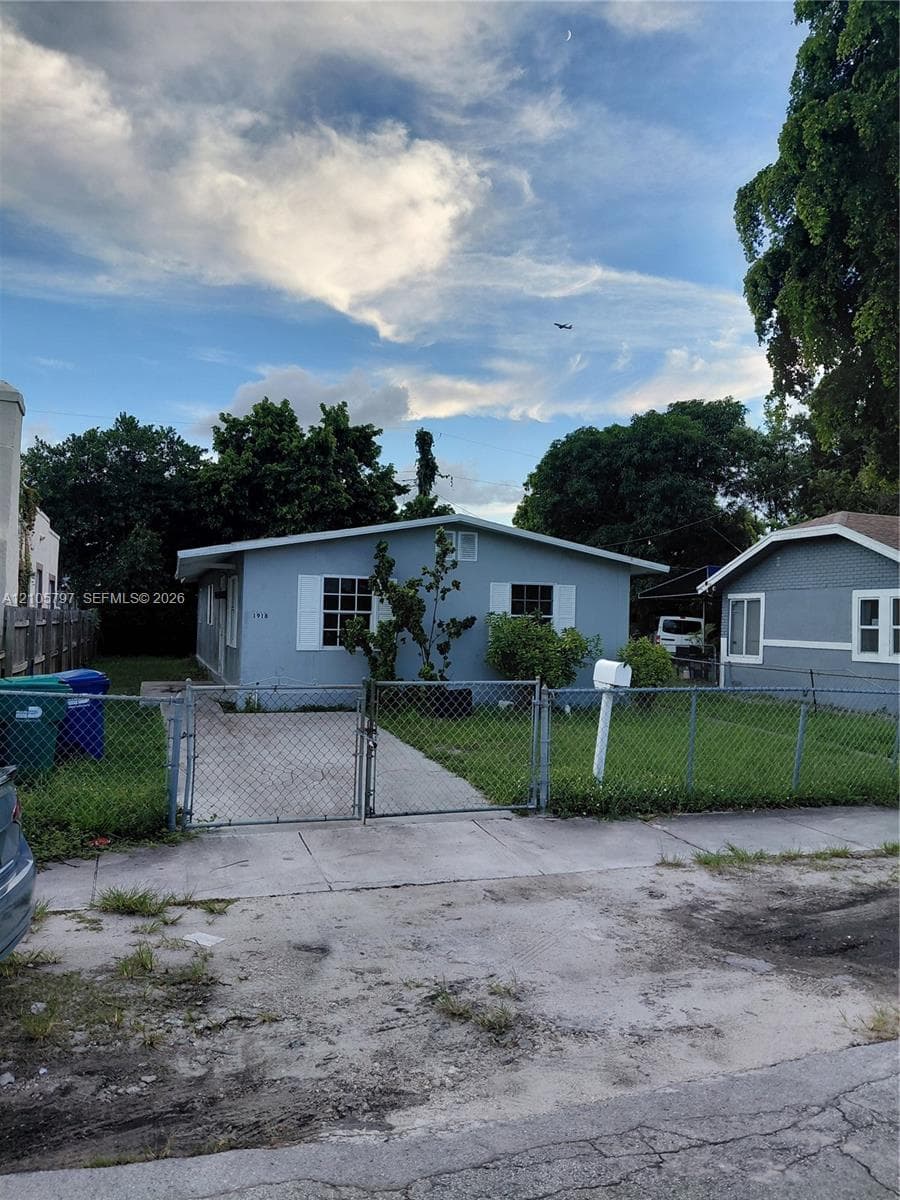 Single-story exterior featuring light-toned lap siding, a metal chain-link perimeter fence, concrete driveway, and white window shutters