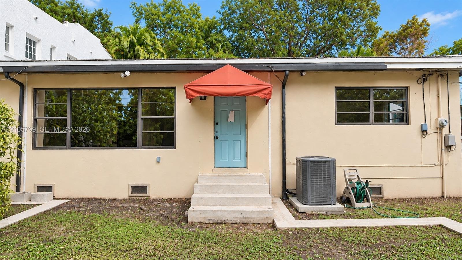 Stucco exterior featuring horizontal casement windows, a central entry with a fabric awning, concrete step access, and a dedicated outdoor heating and cooling unit