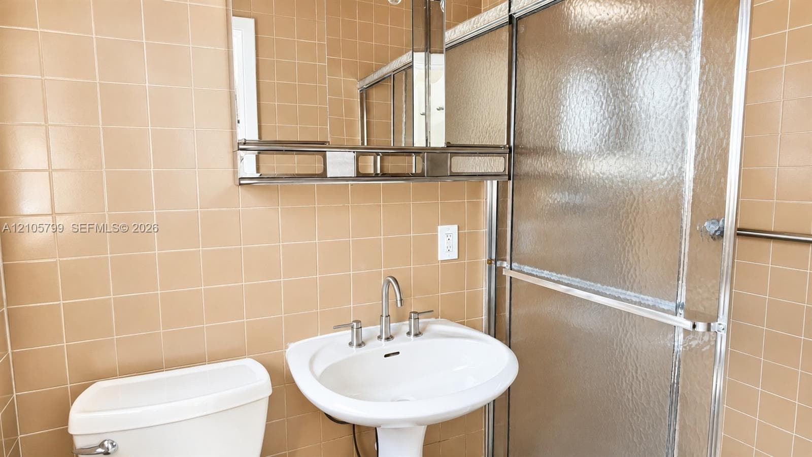 Wall-mounted medicine cabinet with integrated shelving, pedestal sink, metallic widespread faucet, and shower enclosure with textured glass doors, set against floor-to-ceiling tiled walls