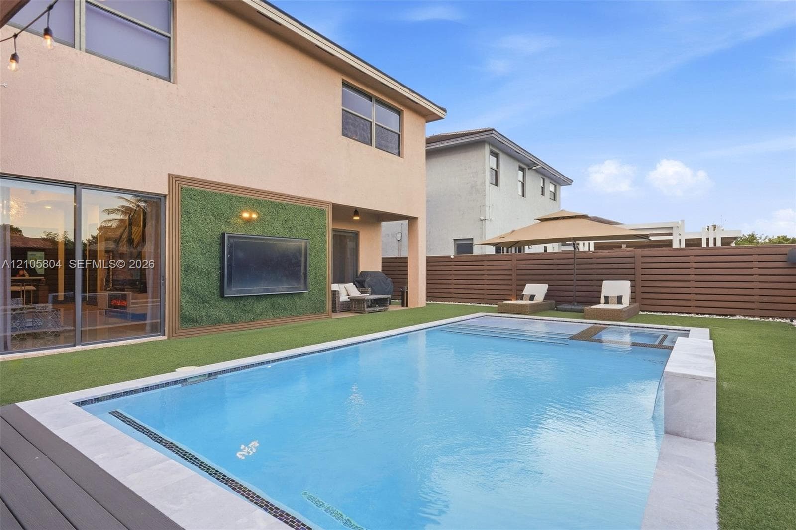 Backyard featuring an in-ground swimming pool with a built-in spa, horizontal wood-slat privacy fencing, artificial turf lawn, and an exterior wall finished with a decorative green panel