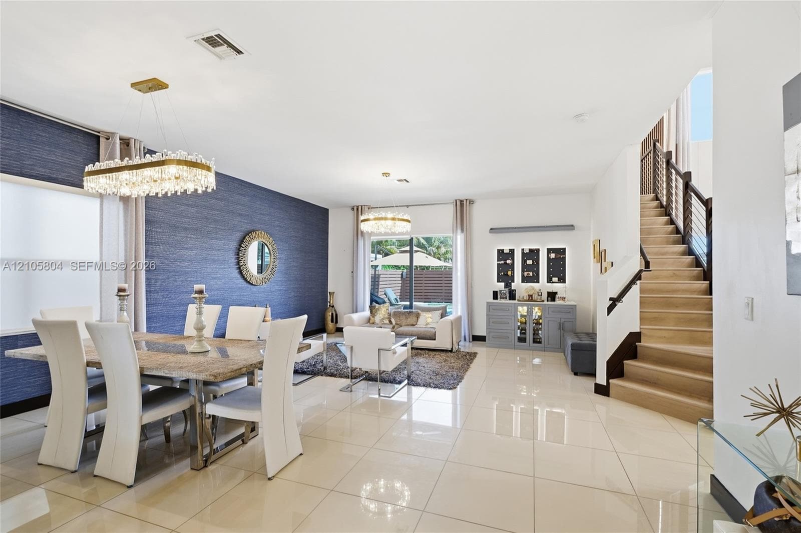 Open-concept interior featuring oversized tile flooring, a blue textured accent wall, dual crystal chandeliers, and a staircase with wood-tread stairs and a metal cable railing system