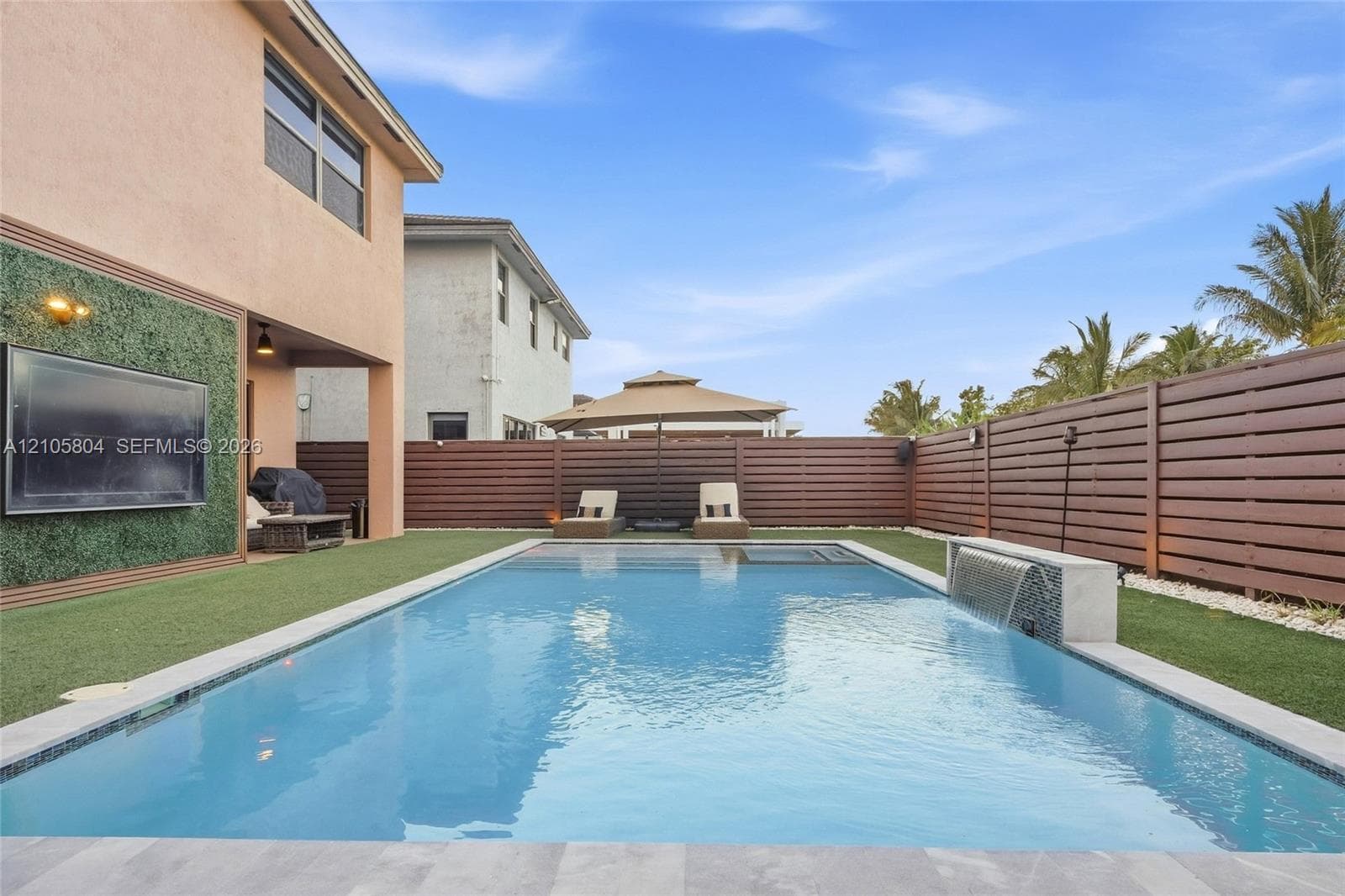 Backyard featuring a rectangular swimming pool with a built-in water feature, horizontal wood-slat privacy fencing, artificial turf lawn, and a wall-mounted exterior television