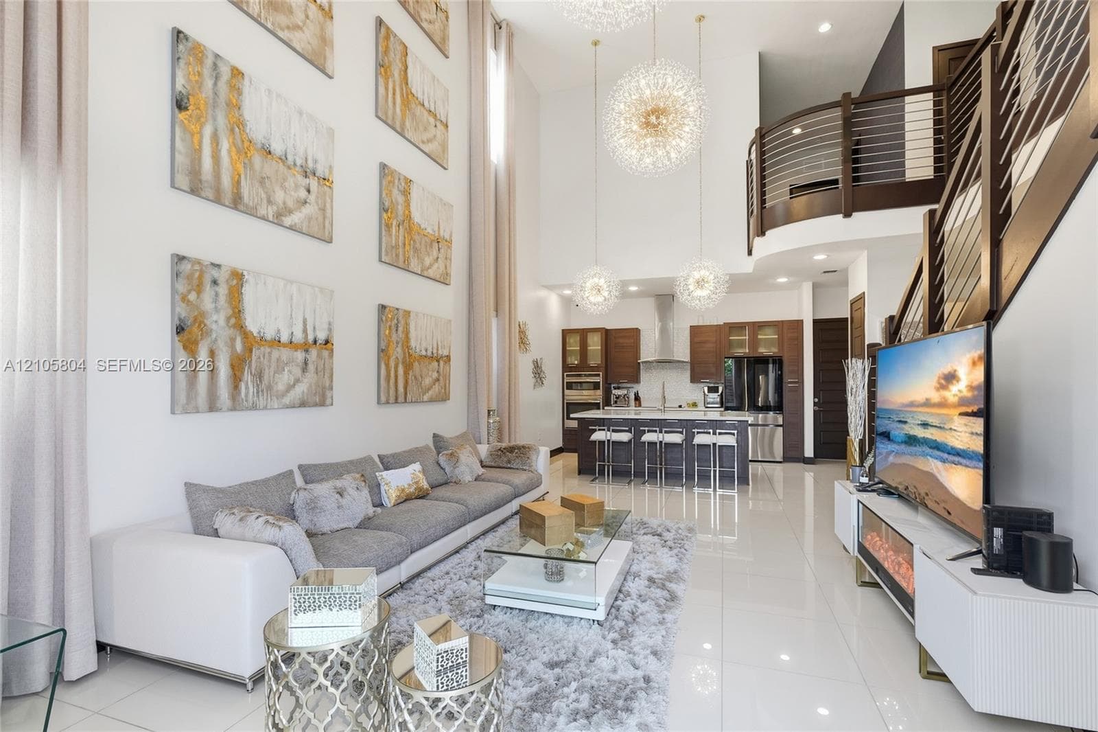 Double-height ceilings, porcelain tile flooring, kitchen featuring custom wood cabinetry and stainless steel appliances, tiered pendant lighting, and metal railed mezzanine overlook