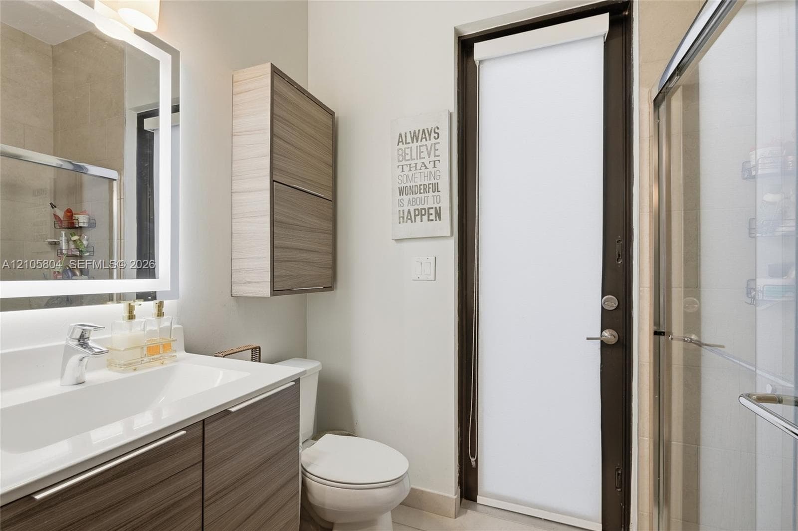 Bathroom featuring a vanity with integrated sink, wood-finish cabinetry, a backlit mirror, a wall-mounted storage cabinet, and a glass-enclosed shower