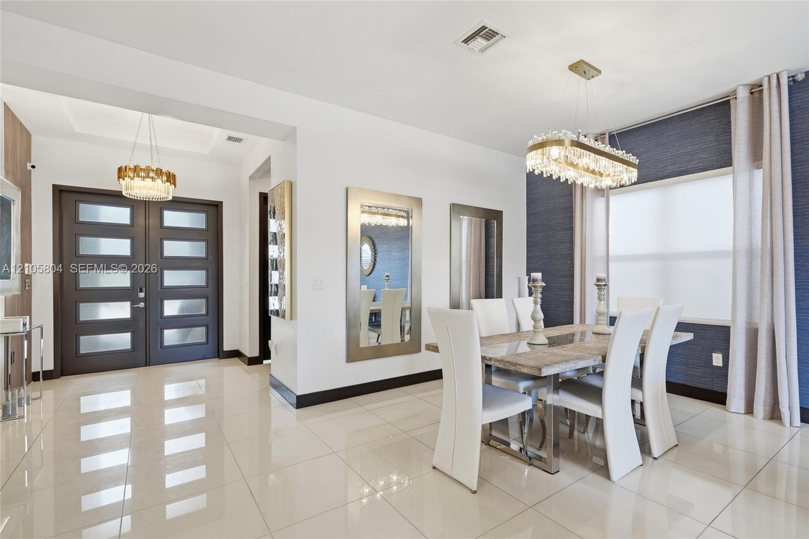 Dining area featuring large-format tile flooring, textured wall covering, custom linear crystal chandelier, and double front entry doors with frosted glass panels