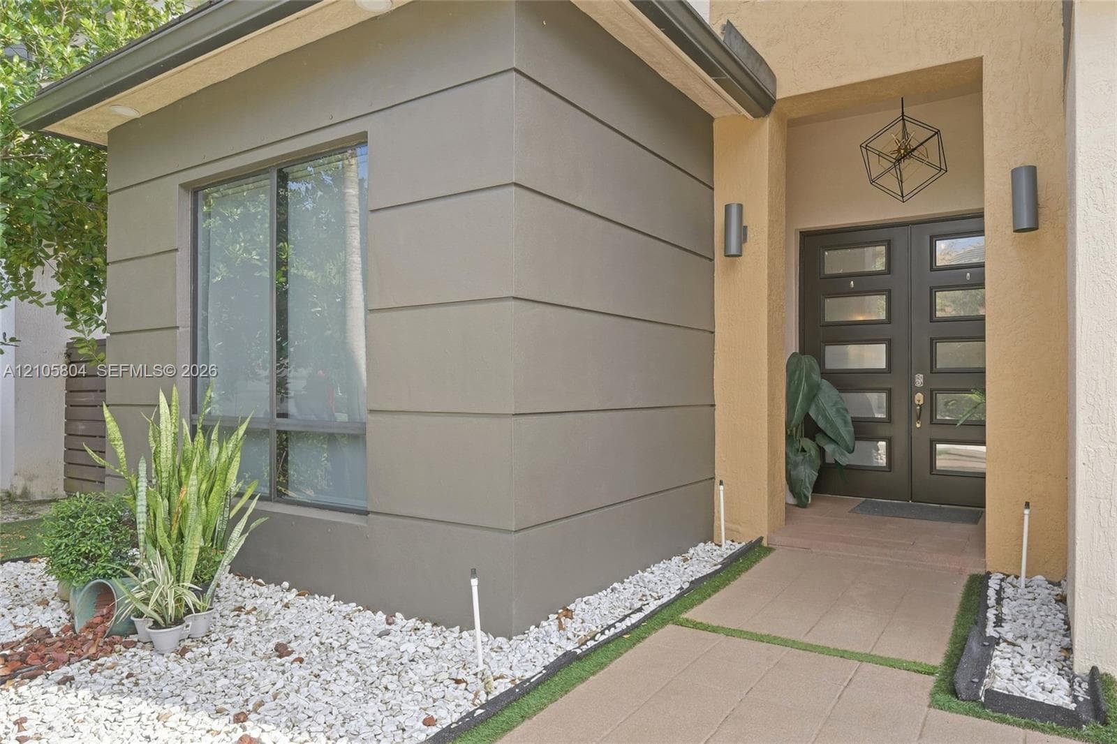 Modern exterior facade featuring smooth stucco finishes, horizontal architectural detailing, double-entry door with glass panels, geometric metal pendant lighting, and a paved stone walkway