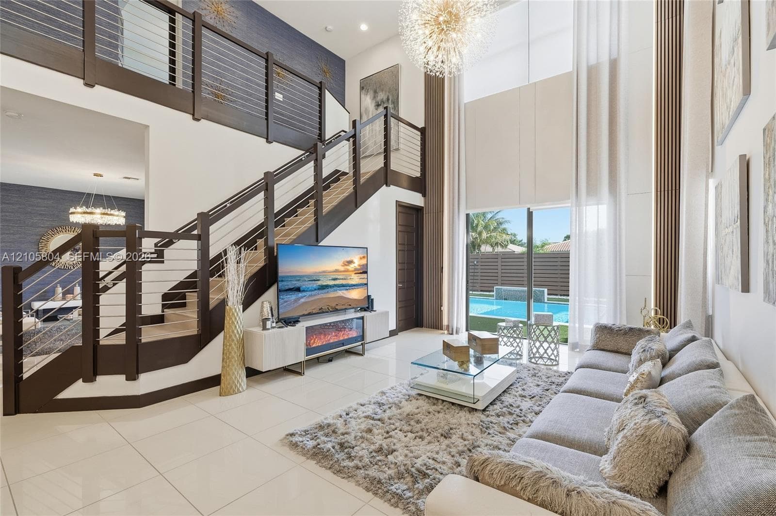 Double-height living area featuring large-format tile flooring, a custom staircase with dark wood-finish treads and a horizontal cable railing system, a recessed electric fireplace, and floor-to-ceiling windows
