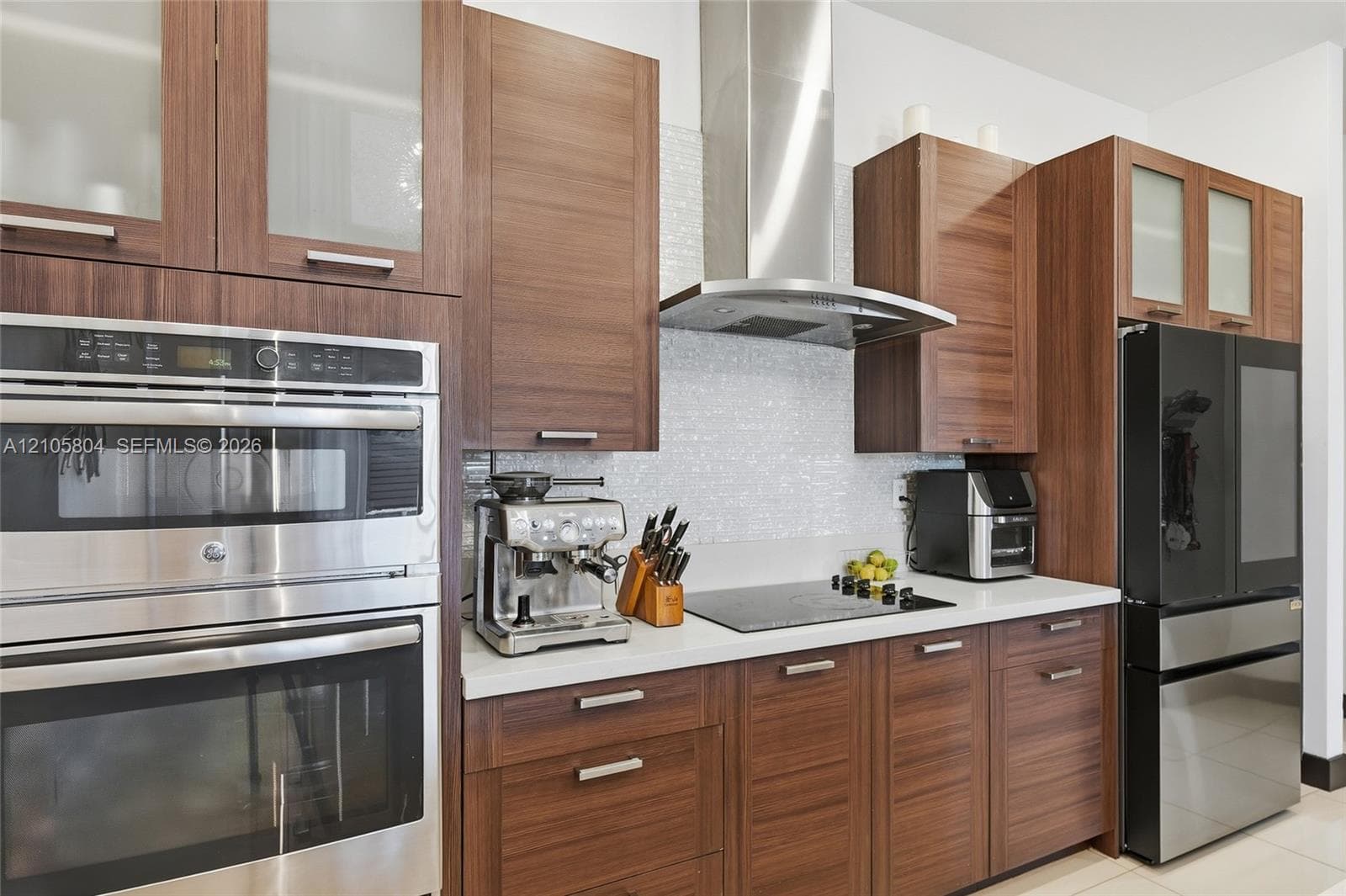 Kitchen featuring wood-finish cabinetry, stone-surface countertops, textured tile backsplash, stainless steel double wall ovens, and a stainless steel range hood