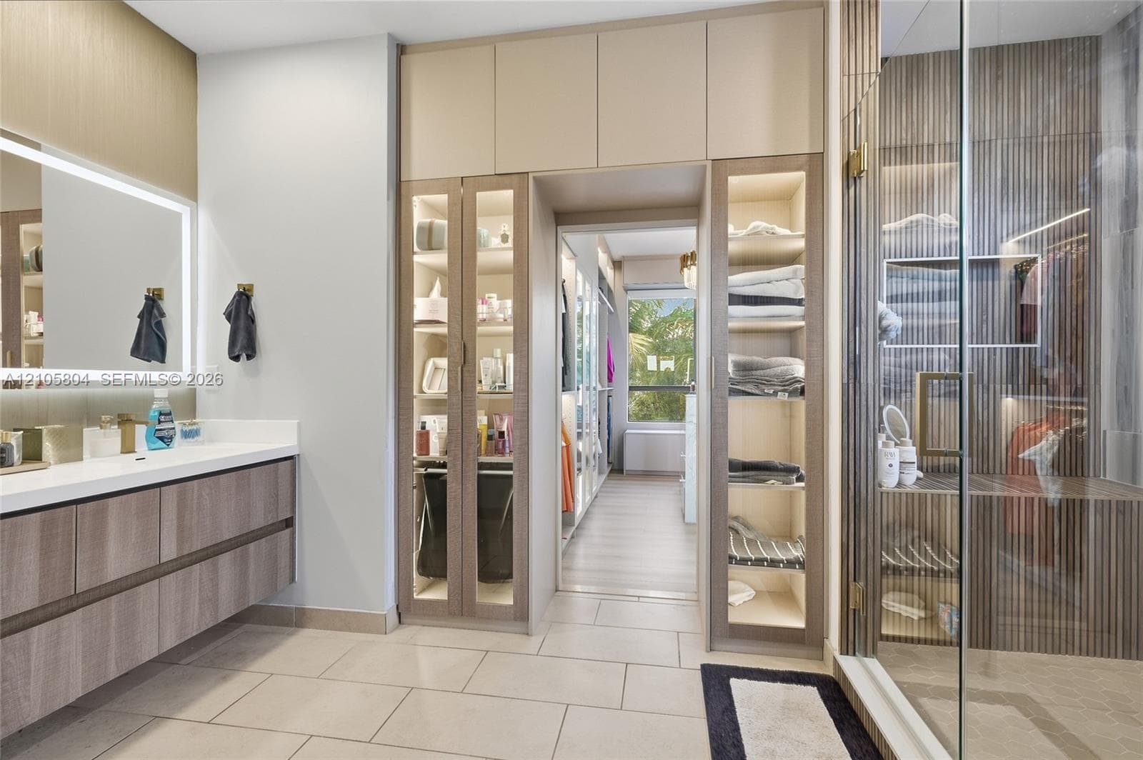 Floating vanity with stone countertop and backlit mirror, floor-to-ceiling built-in cabinetry featuring glass-front display panels, and a walk-in shower with vertical wood-finish wall paneling