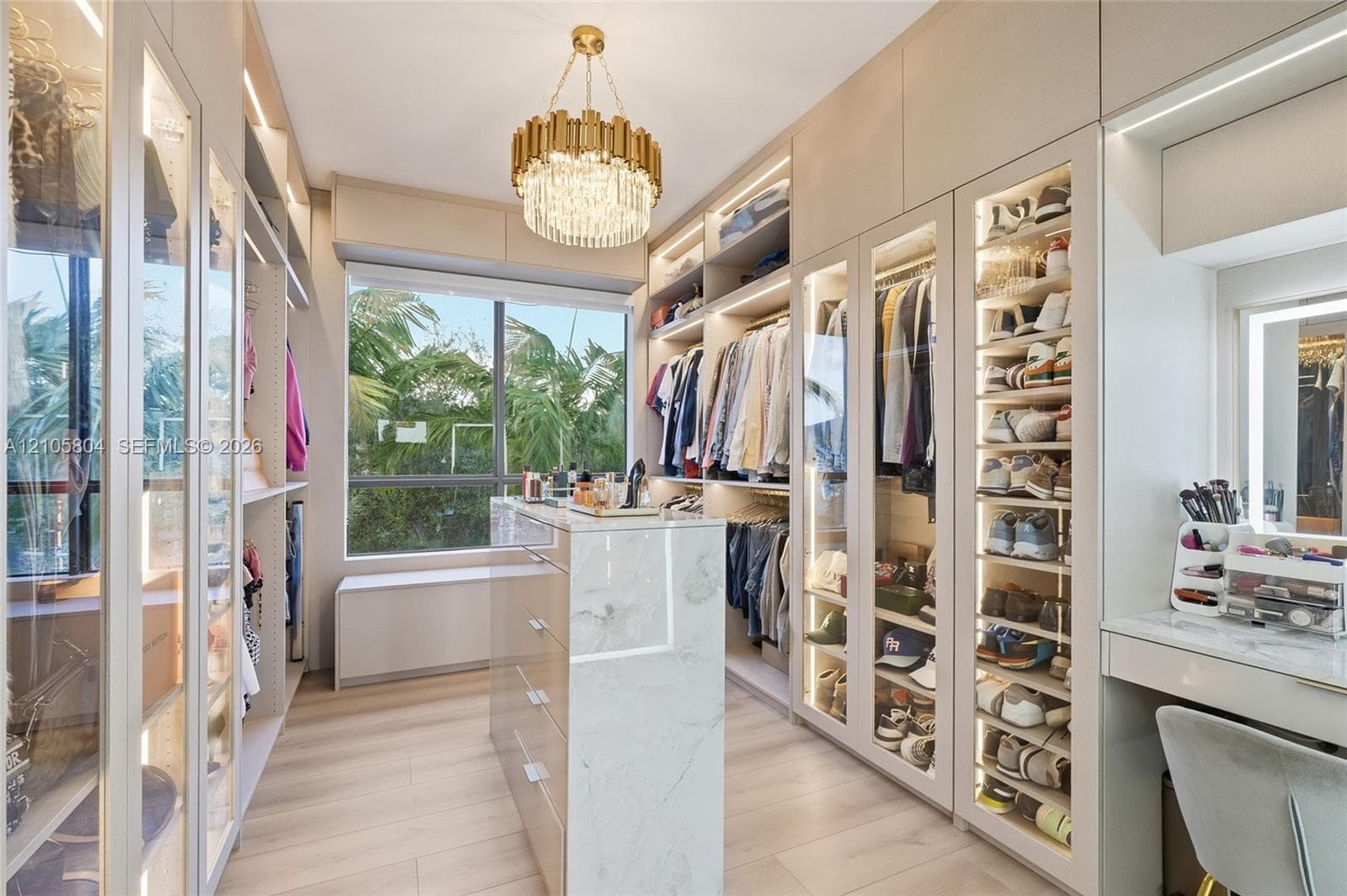 Custom walk-in closet featuring floor-to-ceiling glass-front cabinetry, integrated LED lighting, stone-finish center island, light wood-finish flooring, and a dedicated vanity station