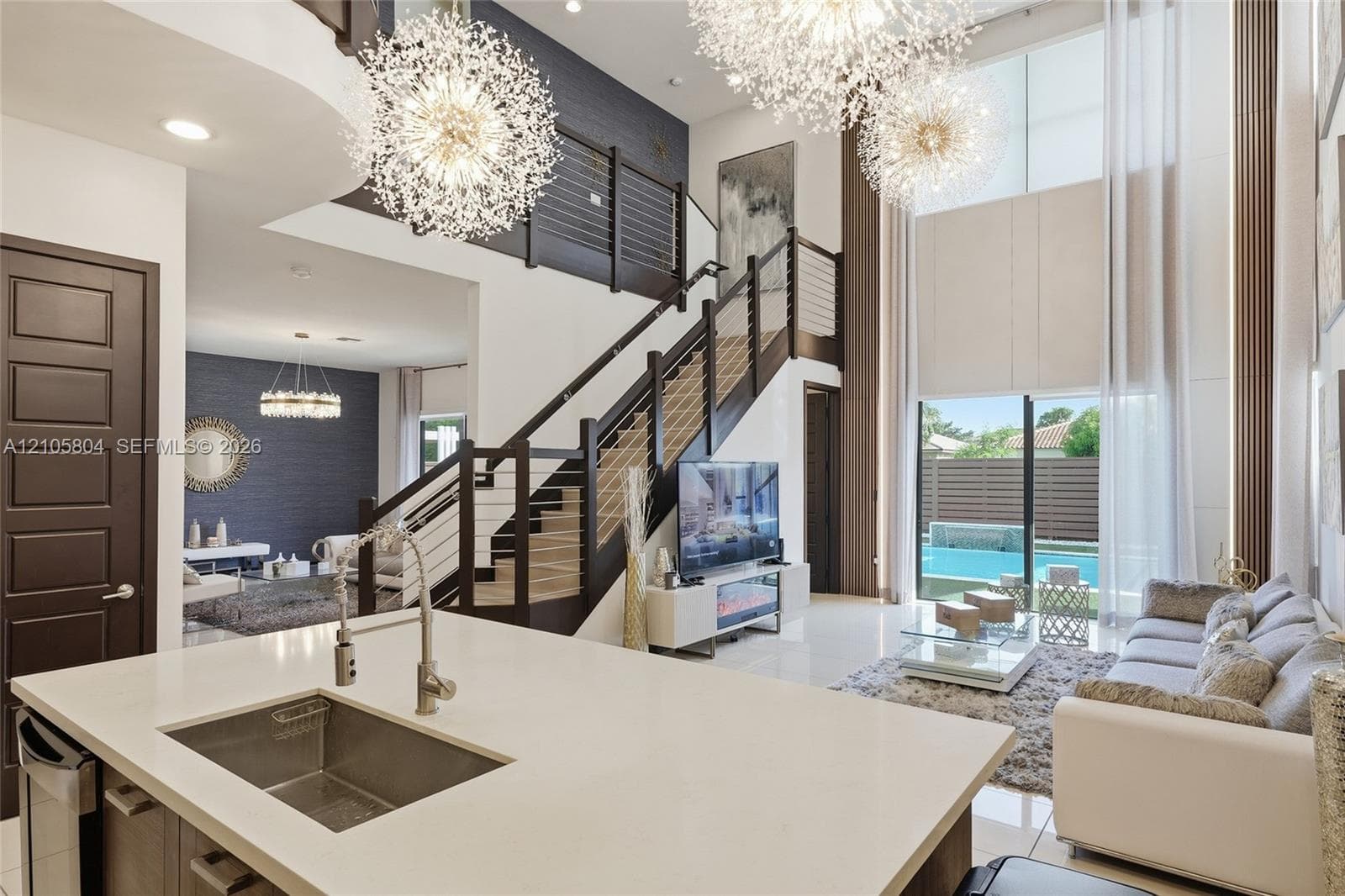 Double-height ceiling, open-plan interior featuring a floating staircase with cable railing, expansive floor-to-ceiling windows, dual sputnik-style chandeliers, and a kitchen island with a stone-finish countertop