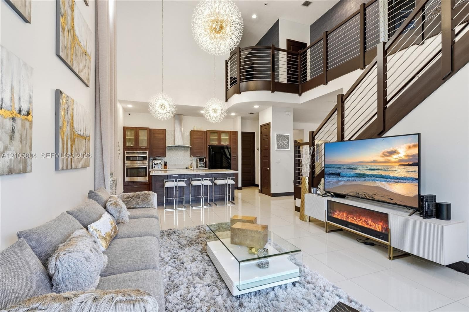 Double-height ceilings featuring tiered spherical pendant lighting, polished stone-finish flooring, and a curved staircase with dark-stained wood handrails and horizontal metal balusters