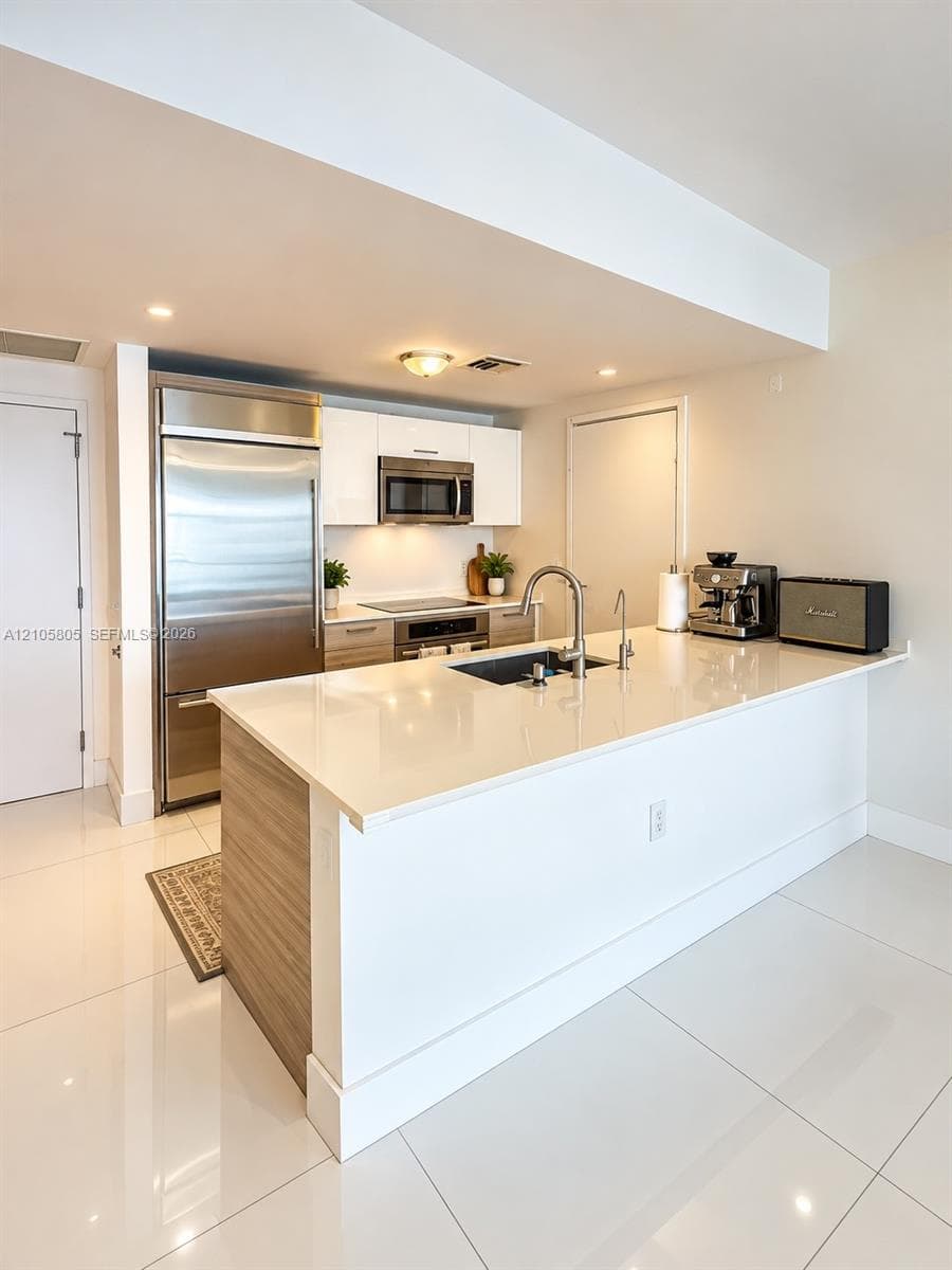 Kitchen featuring a quartz countertop island with an integrated stainless steel undermount sink, flat-panel cabinetry, stainless steel refrigerator, and high-gloss tile flooring