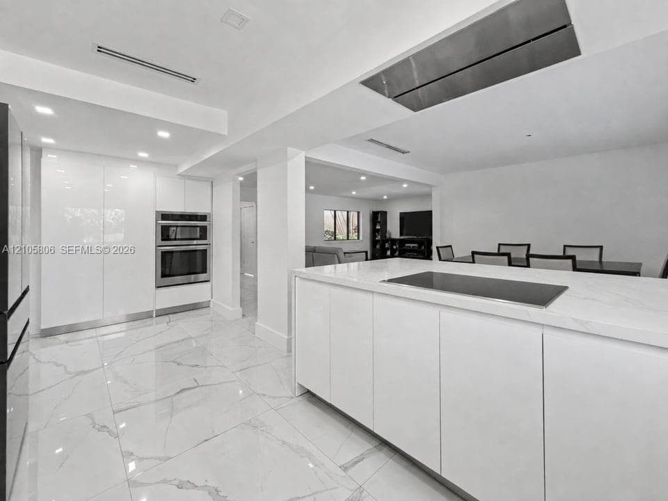 Modern kitchen featuring a central island with an integrated cooktop, stone-finish countertops, high-gloss cabinetry, stainless steel double wall ovens, and large-format tile flooring