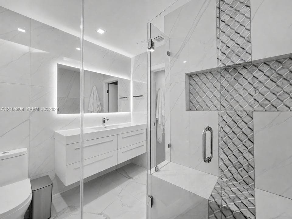 Modern floating vanity with a seamless countertop, integrated basin, and back-lit mirror, complemented by a walk-in glass-enclosed shower featuring detailed stone-tile inlay and a built-in bench