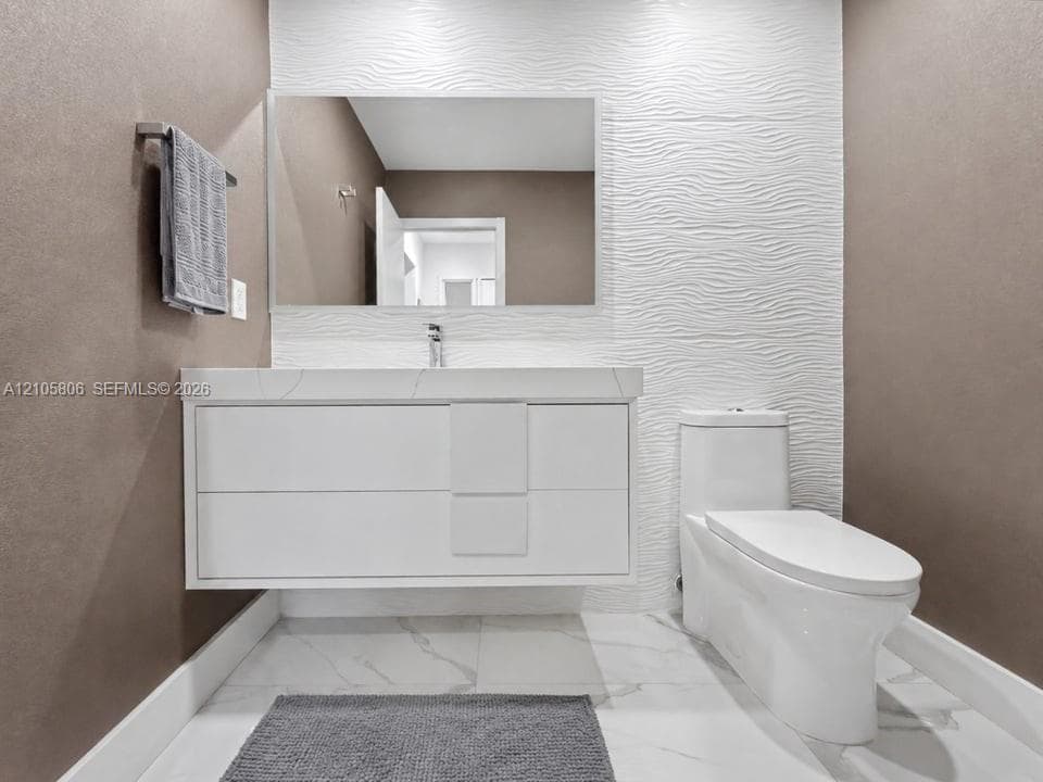 Floating vanity with integrated sink, stone-surface countertop, textured white feature wall, large-format tiled flooring, and dual-flush toilet
