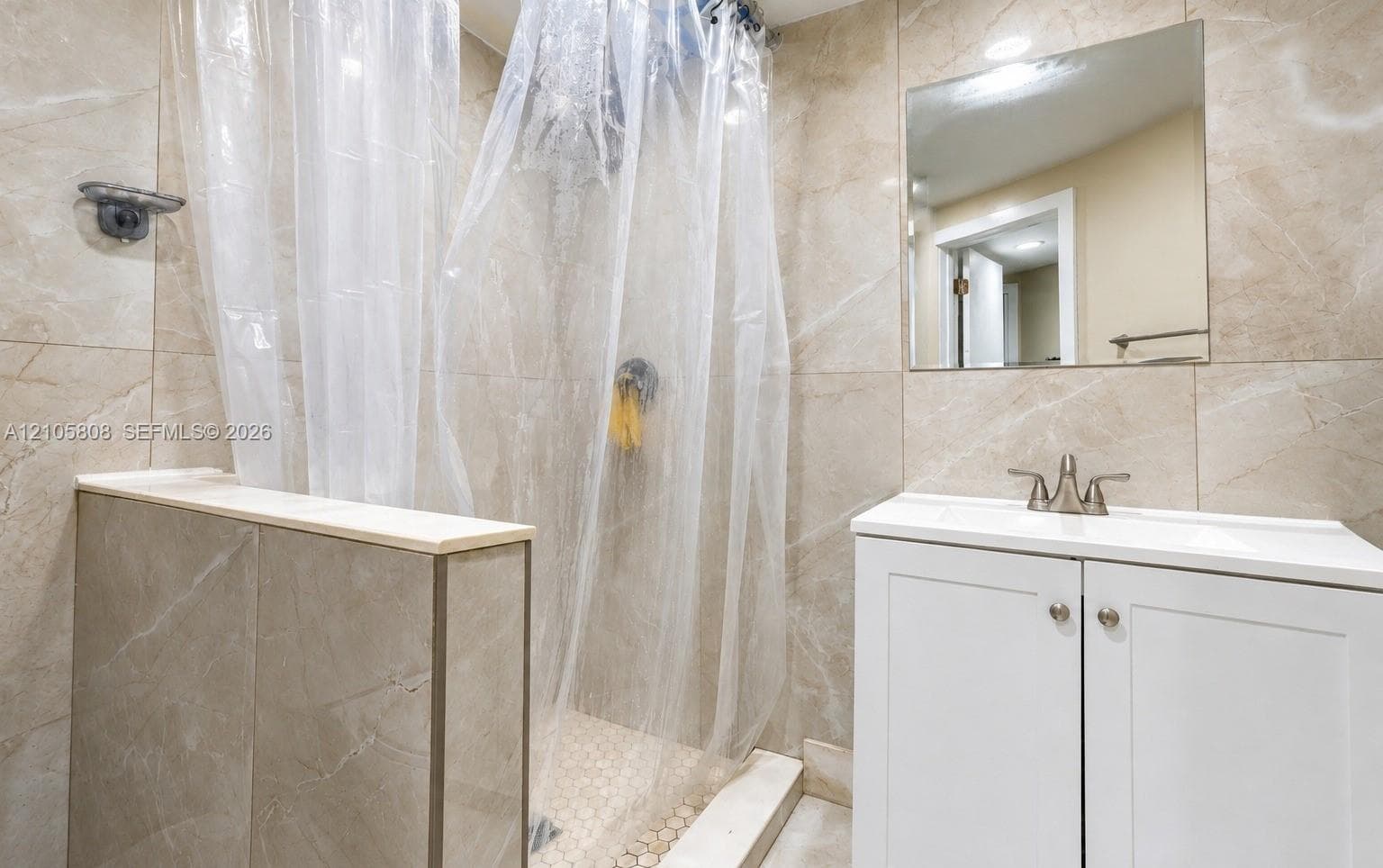 Walk-in shower enclosure featuring floor-to-ceiling neutral tile, white shaker-style vanity cabinet, integrated sink with dual-handle faucet, wall-mounted mirror, and tile flooring