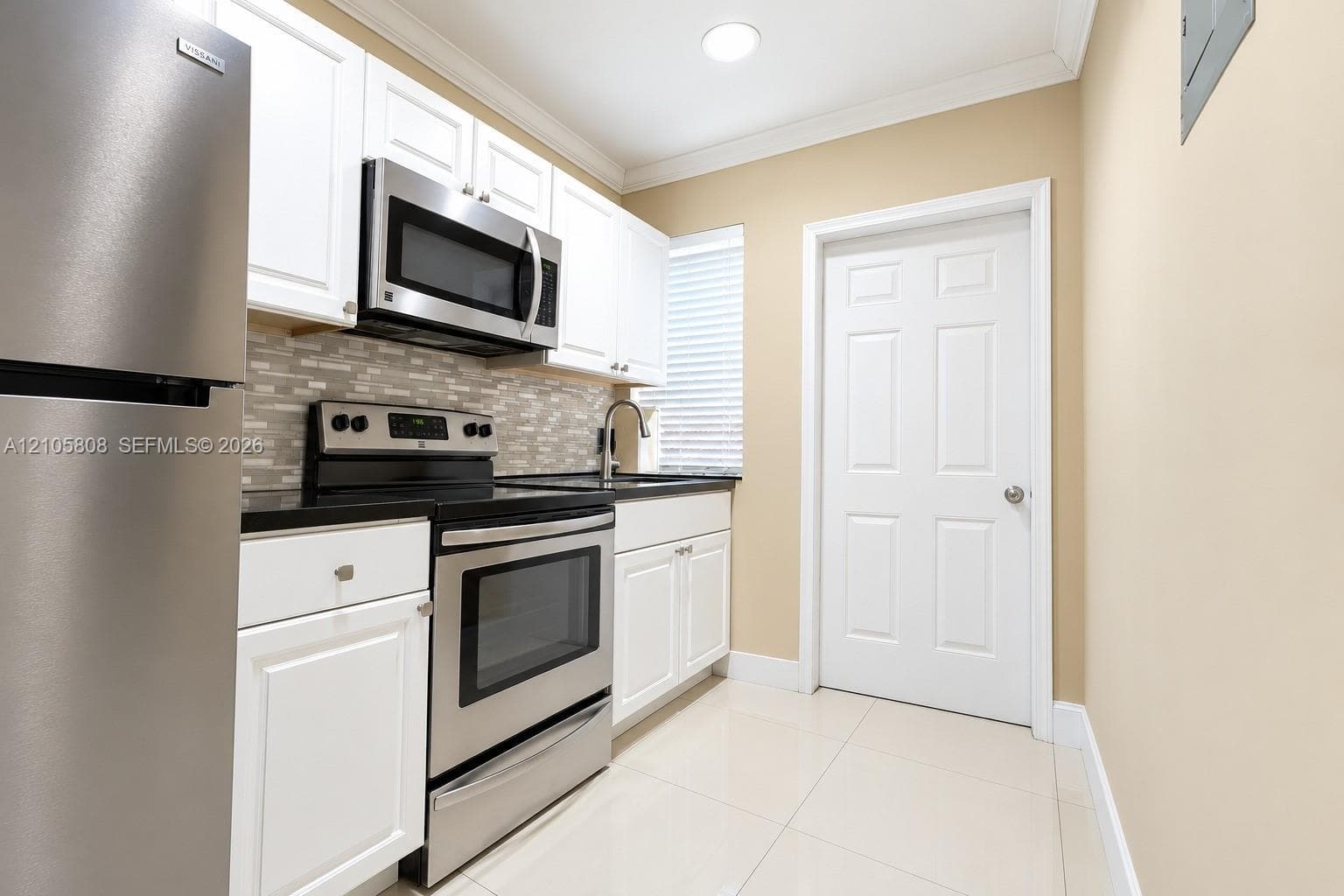 Kitchen featuring white shaker-style cabinetry, stainless steel appliances, dark stone countertops, a mosaic tile backsplash, and light-toned tile flooring