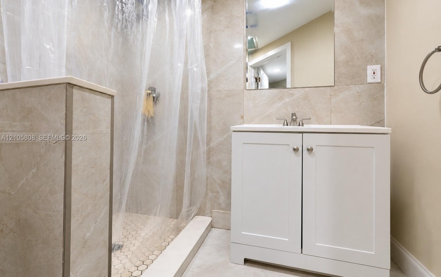 White vanity cabinet with a stone countertop, wall-mounted mirror, walk-in shower with large-format tile surround, and hexagonal tile flooring