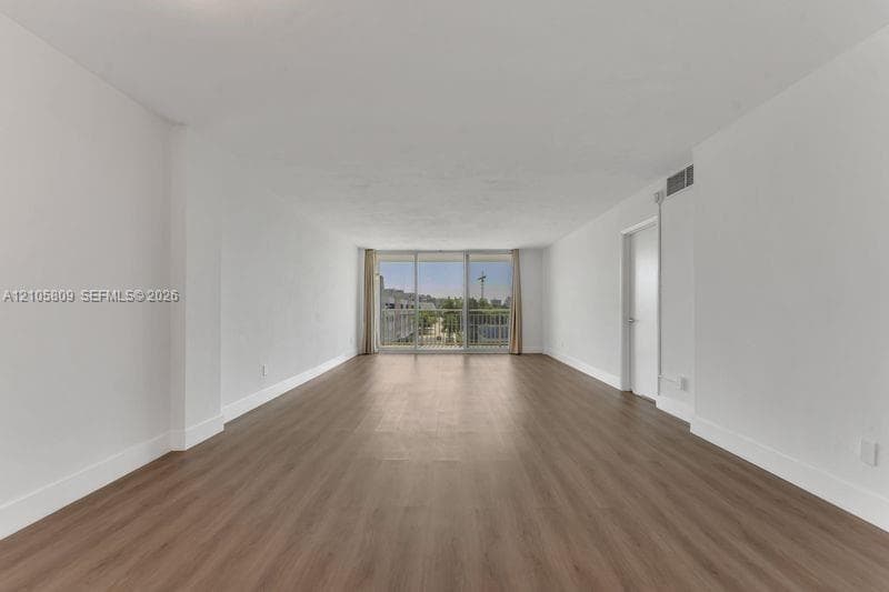 Open interior space featuring wood-finish flooring, neutral walls, and a floor-to-ceiling glass sliding door providing access to a metal-railing balcony