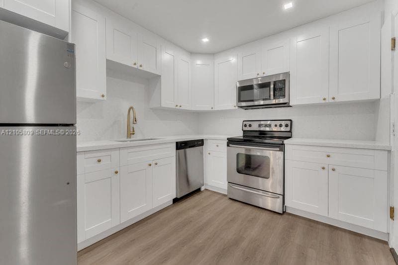 Kitchen featuring white Shaker-style cabinetry, stainless steel appliances, wood-finish flooring, solid-surface countertops, and a brass-tone gooseneck faucet