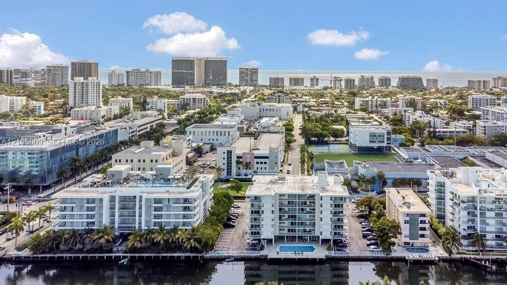 Aerial perspective of waterfront mid-rise buildings featuring a ground-level swimming pool, expansive parking, and proximity to municipal infrastructure and coastal urban landscape