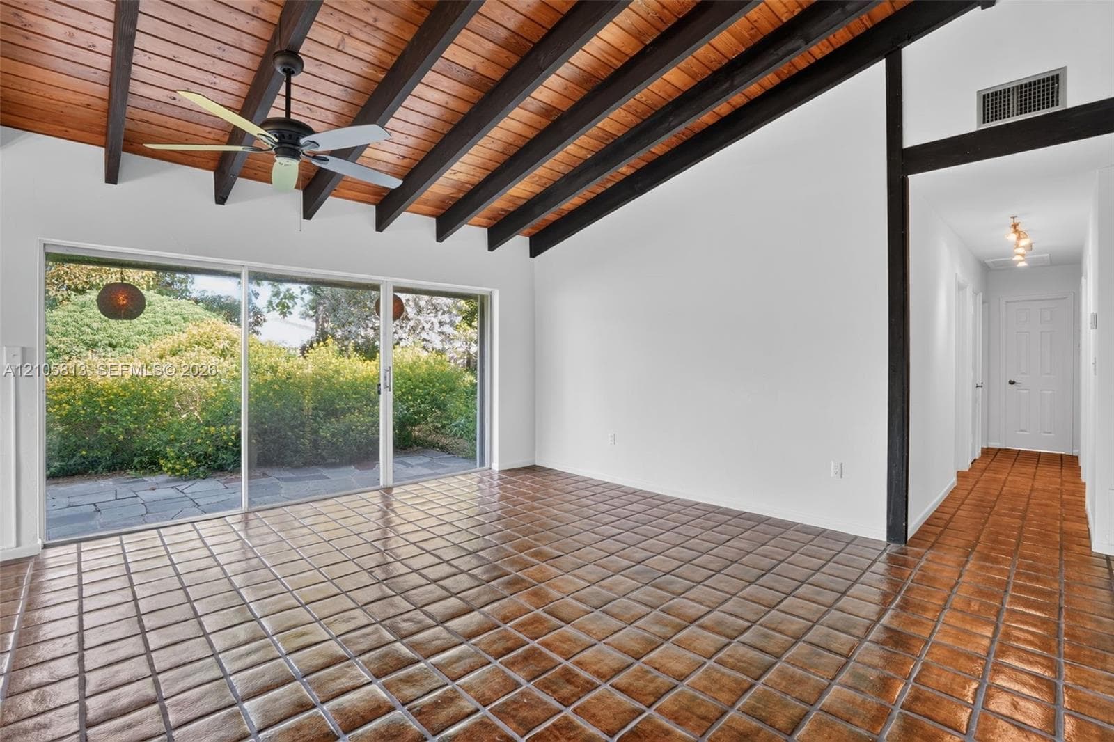 Vaulted ceiling featuring exposed stained wood beams and natural wood-finish planking, complemented by glazed terra-cotta floor tiles, sliding glass doors, and a ceiling fan