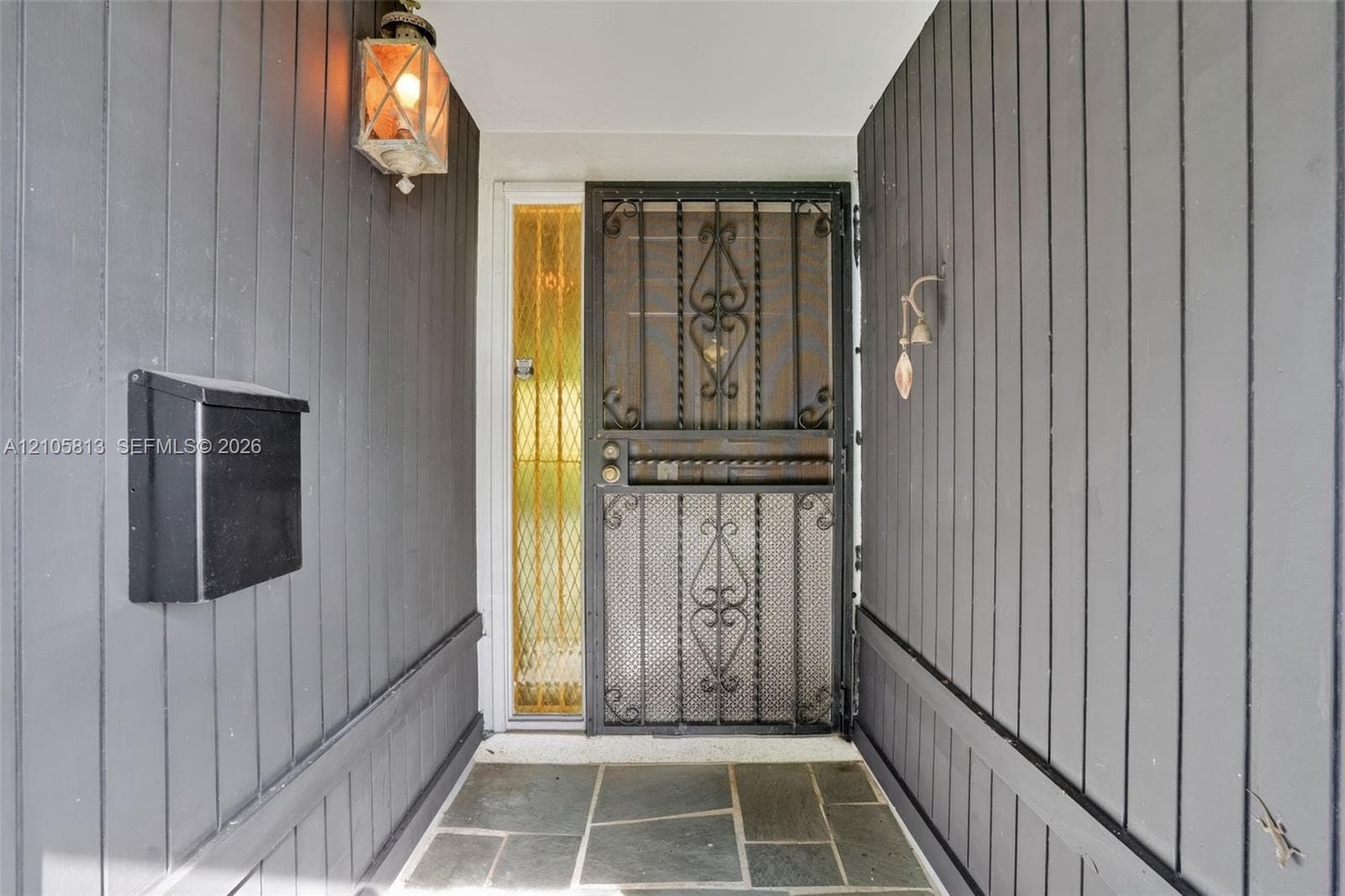Covered entryway featuring vertical dark-tone wood siding, flagstone flooring, a decorative metal security door, a sidelight with textured glass, and a wall-mounted lantern-style fixture