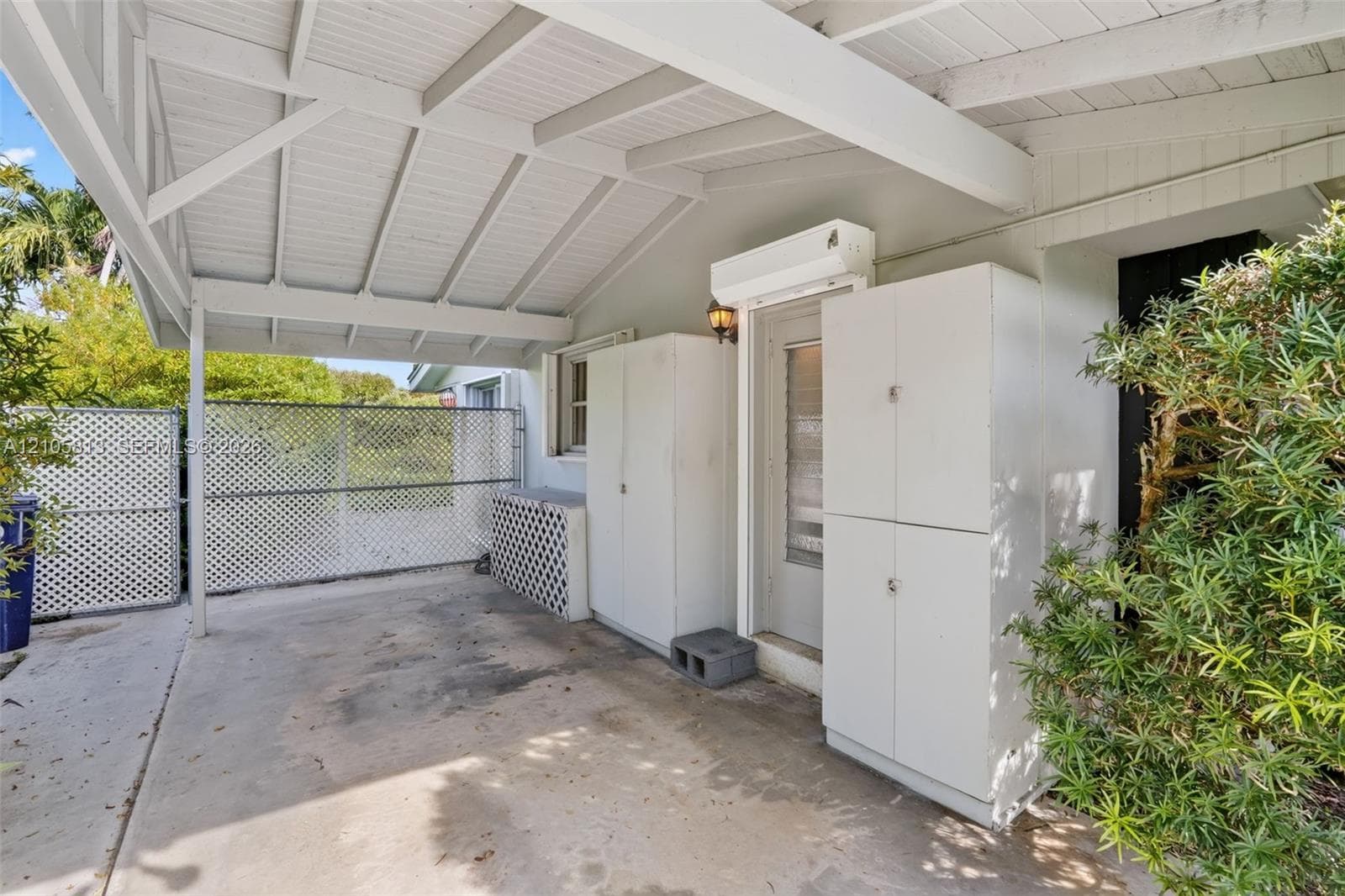 Covered carport featuring a painted wood-beamed ceiling, concrete flooring, exterior storage cabinetry, and perimeter lattice fencing