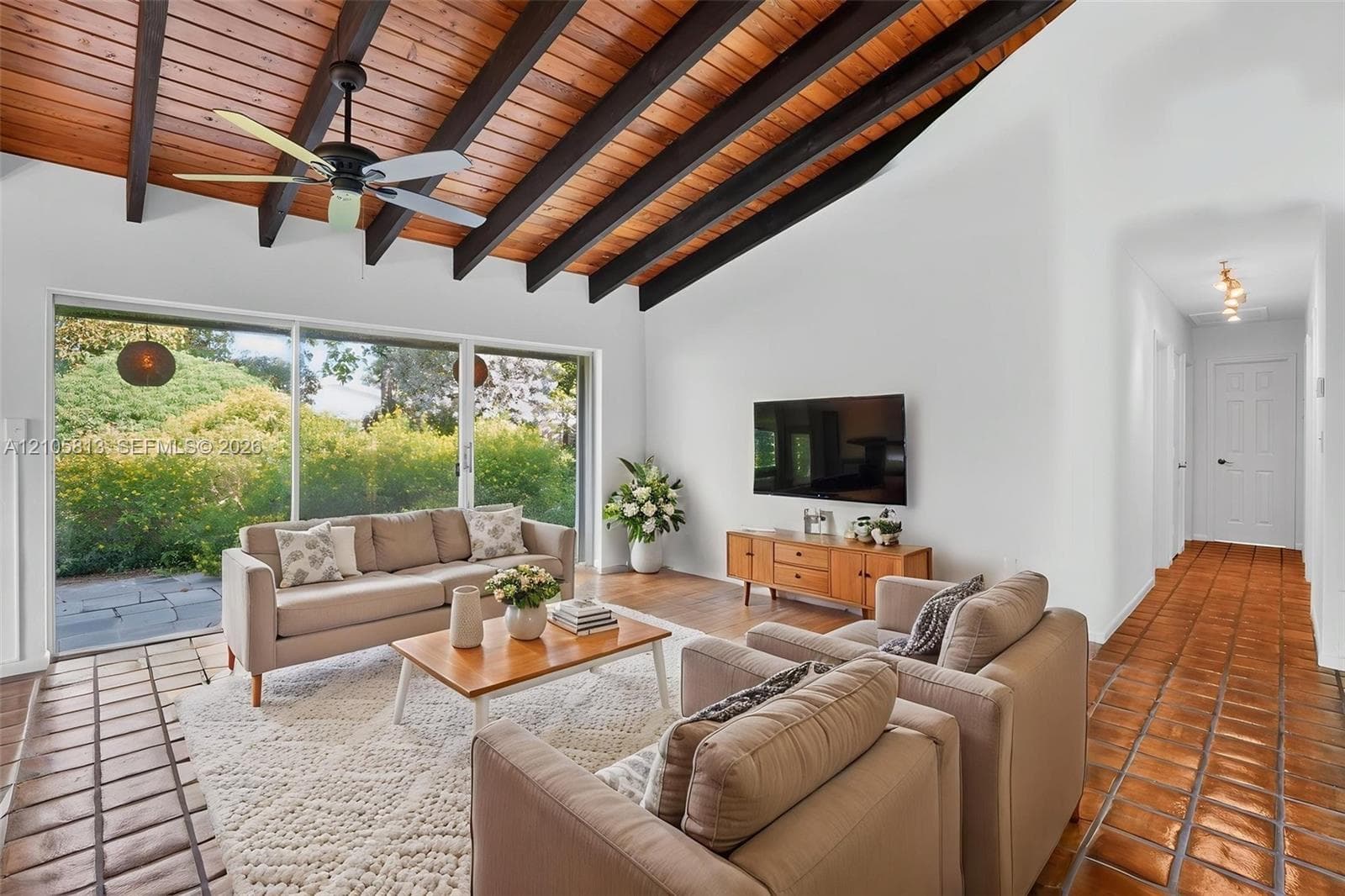 Vaulted ceiling featuring exposed wood beams, stained wood planking, and a central ceiling fan, complemented by extensive floor-to-ceiling glass sliding doors and terracotta tile flooring